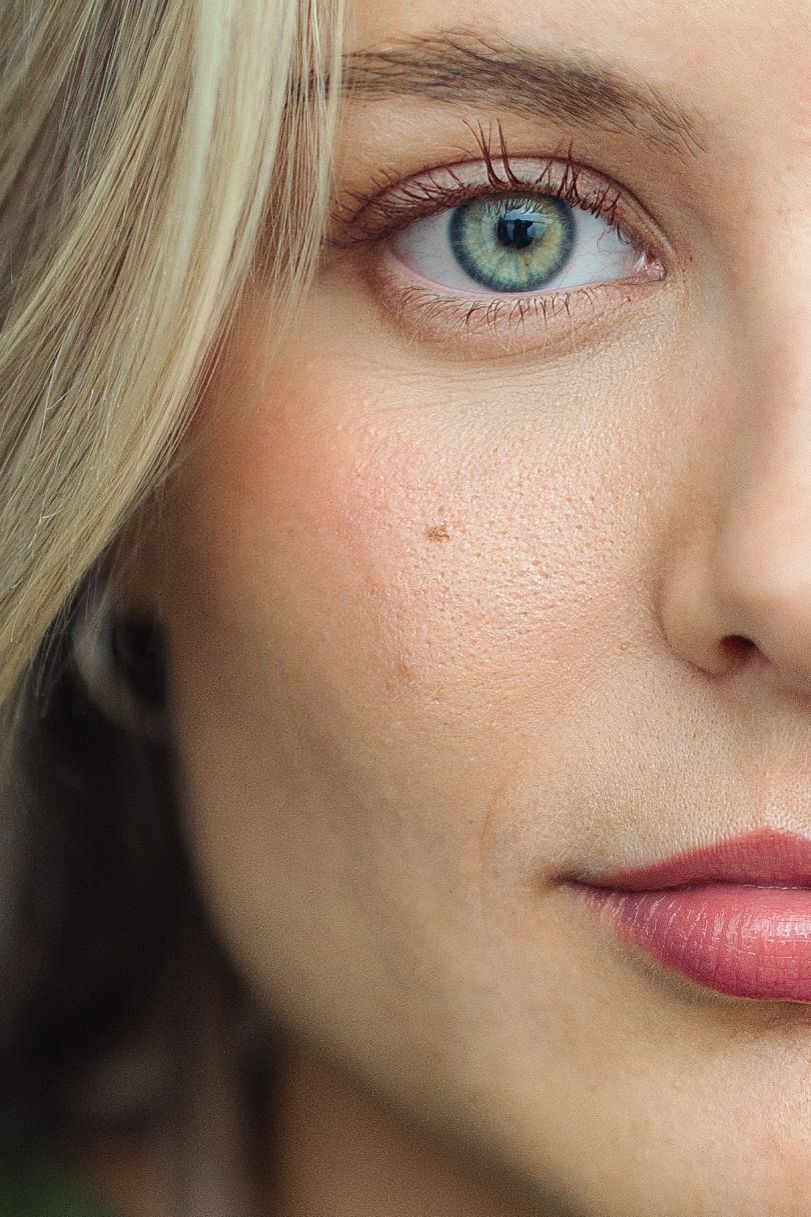 Close-up portrait of a young woman with striking green eyes, blonde hair, fair skin, and a small mole on her cheek.