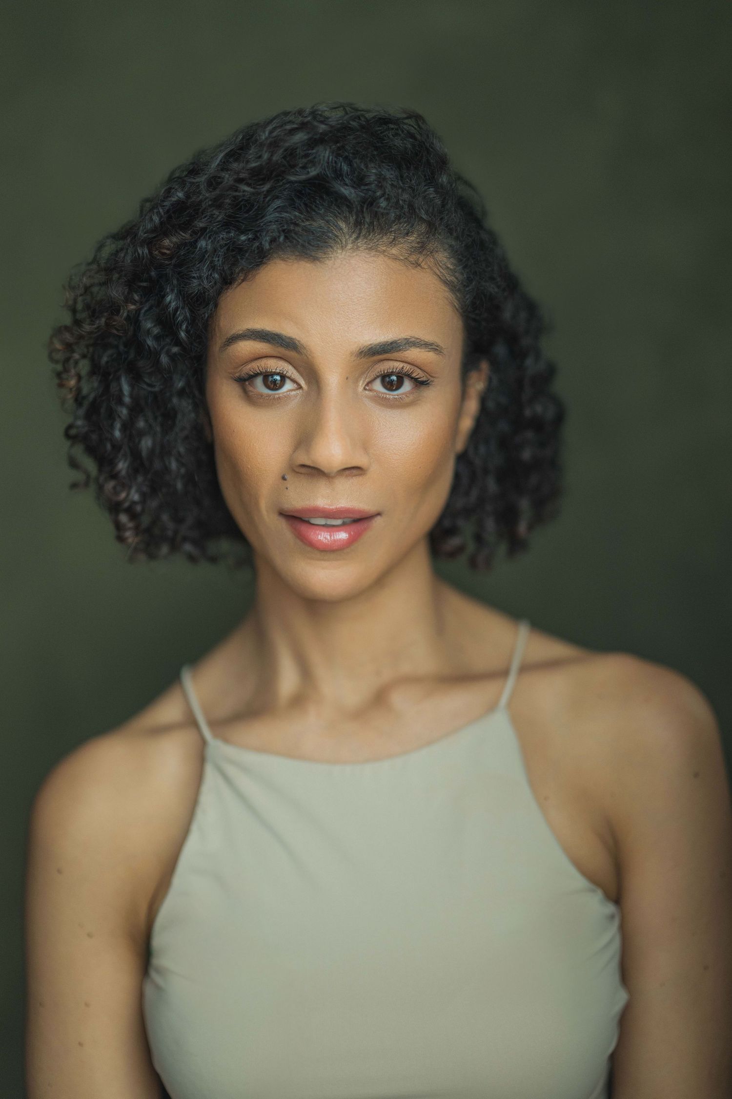 Professional headshot portrait with natural curly hair and sage green tank top against dark green background.