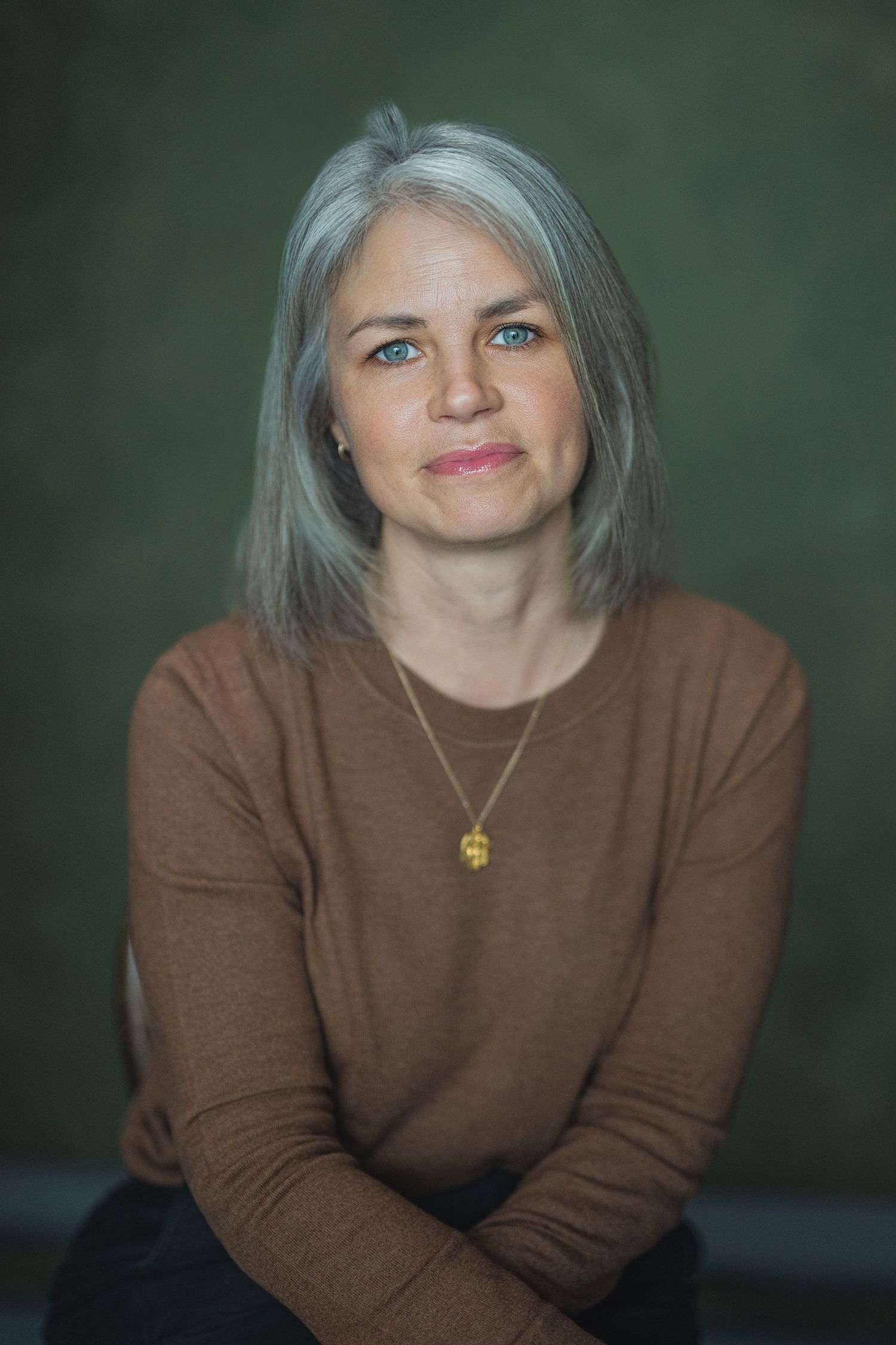 Middle-aged woman with silver hair and blue eyes wearing a brown sweater and gold necklace against a green background.