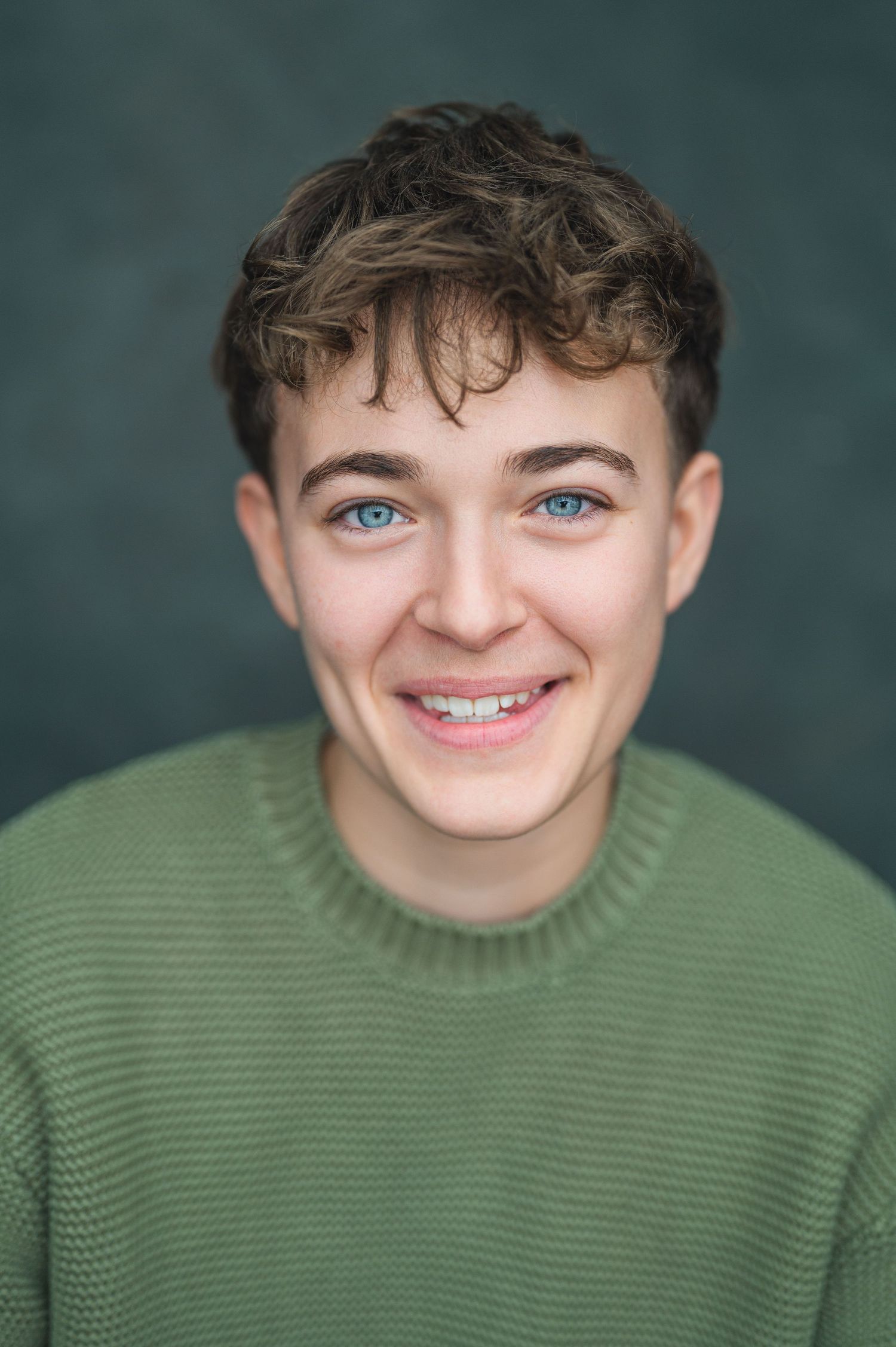 Smiling young man with blue eyes, braces, curly brown hair, wearing a green sweater against a dark background.
