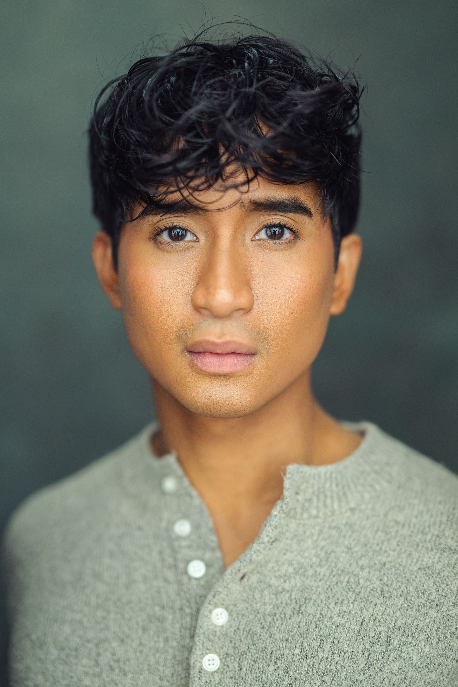 Young man with curly black hair and light eyes wearing a gray henley shirt against a dark background.