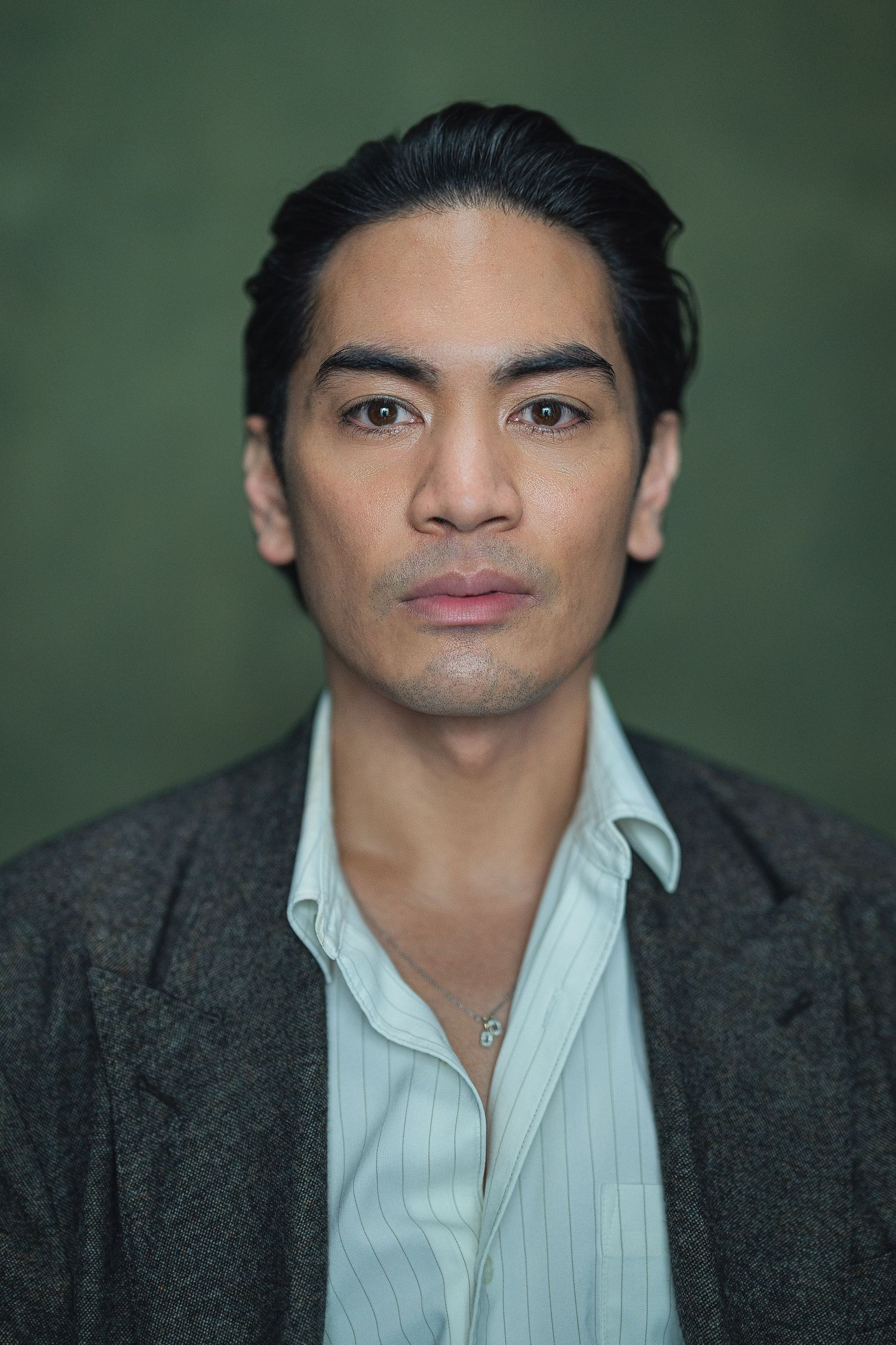 Portrait of a young Asian man in a gray blazer and striped shirt, wearing a necklace, against a dark green background.