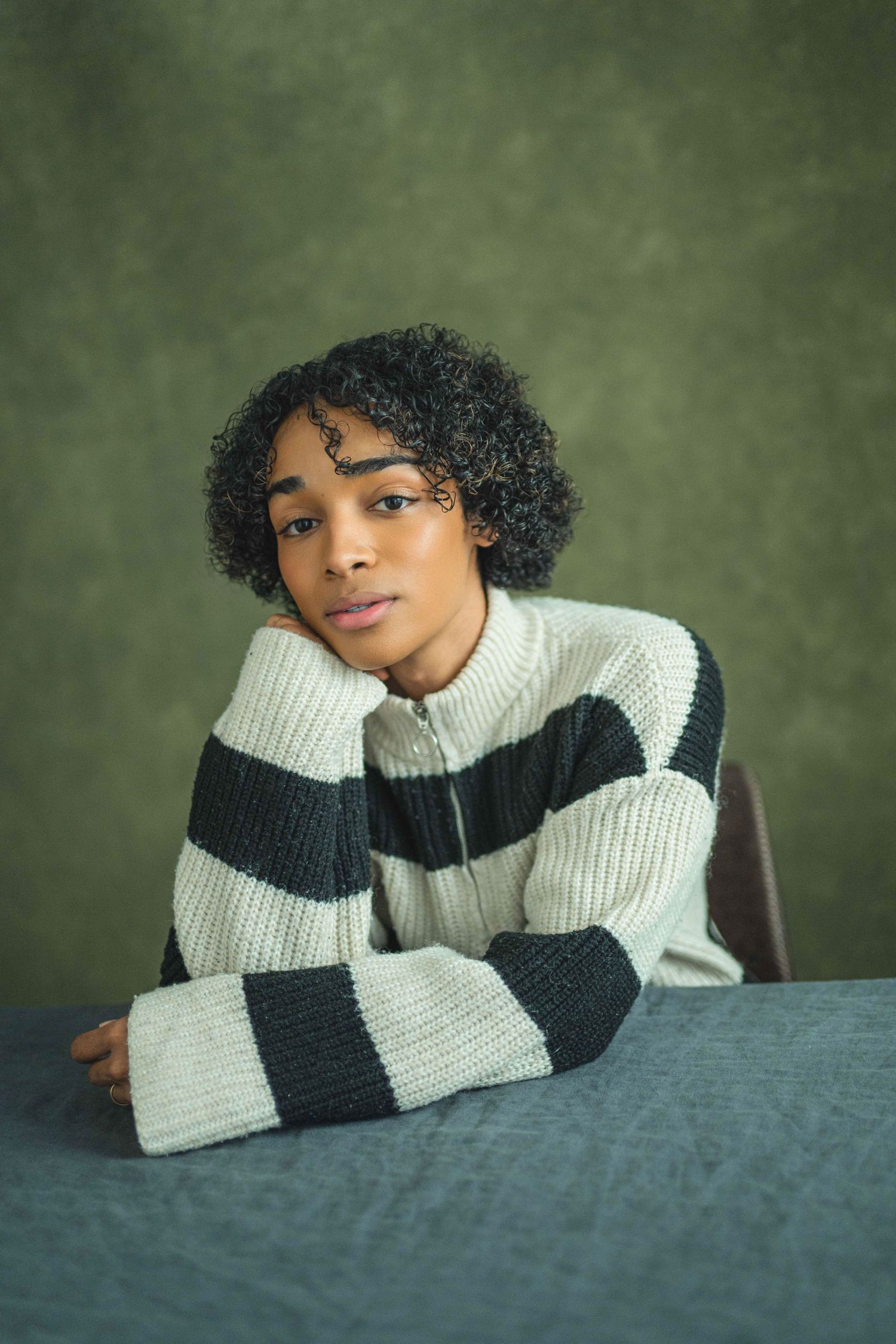 Portrait against green background wearing black and white striped knit sweater while resting on table.
