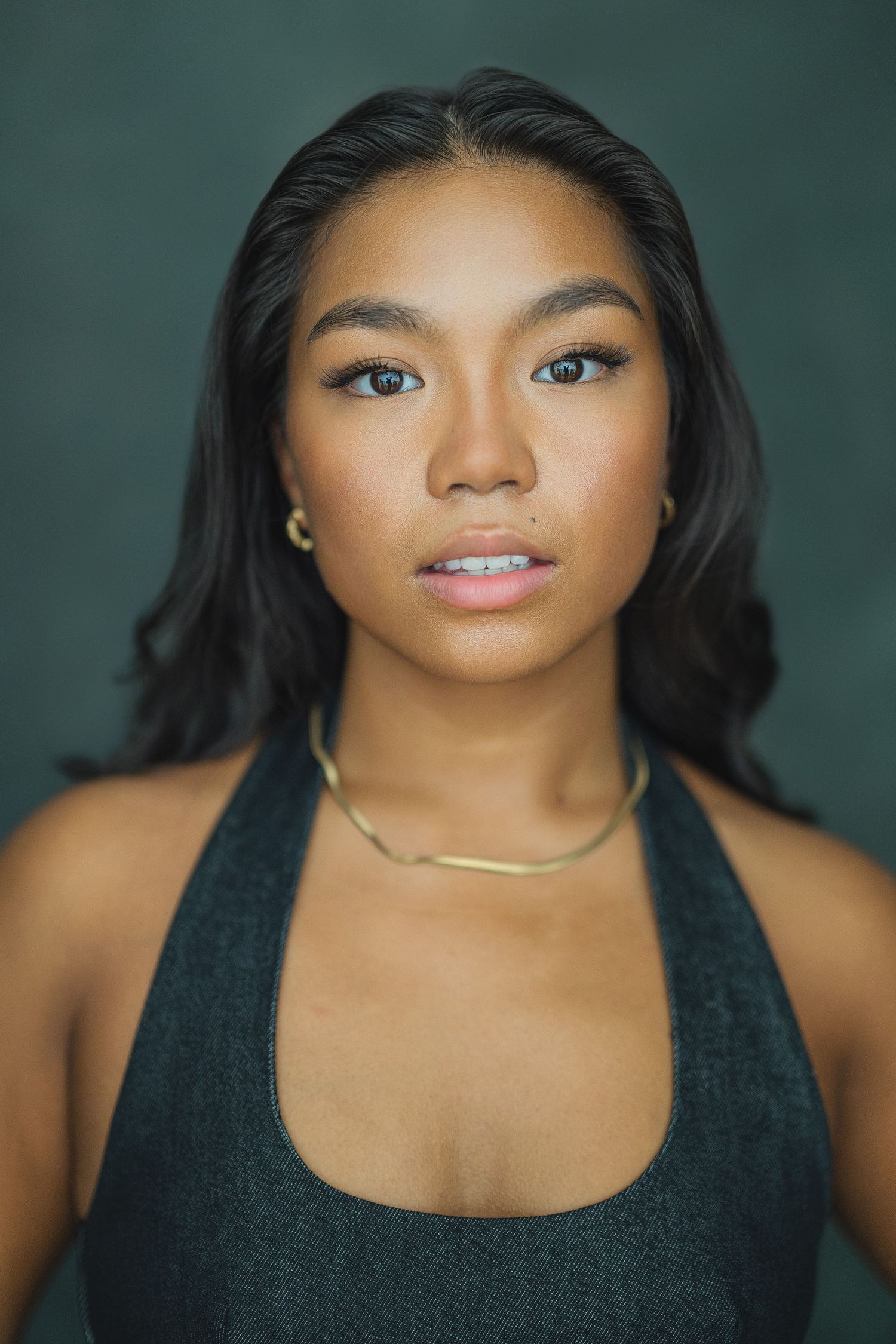 Studio portrait against dark green background wearing black sleeveless top and gold necklace.