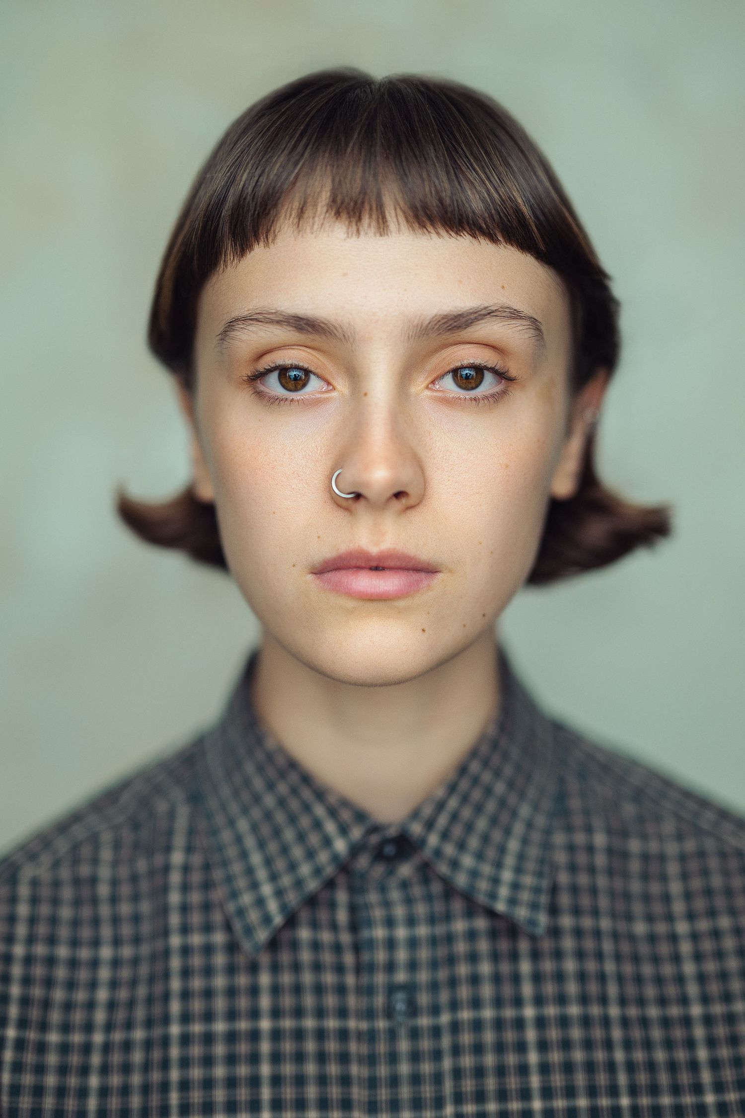 Young person with short dark hair, nose ring, and plaid shirt poses for a portrait against a light green background.