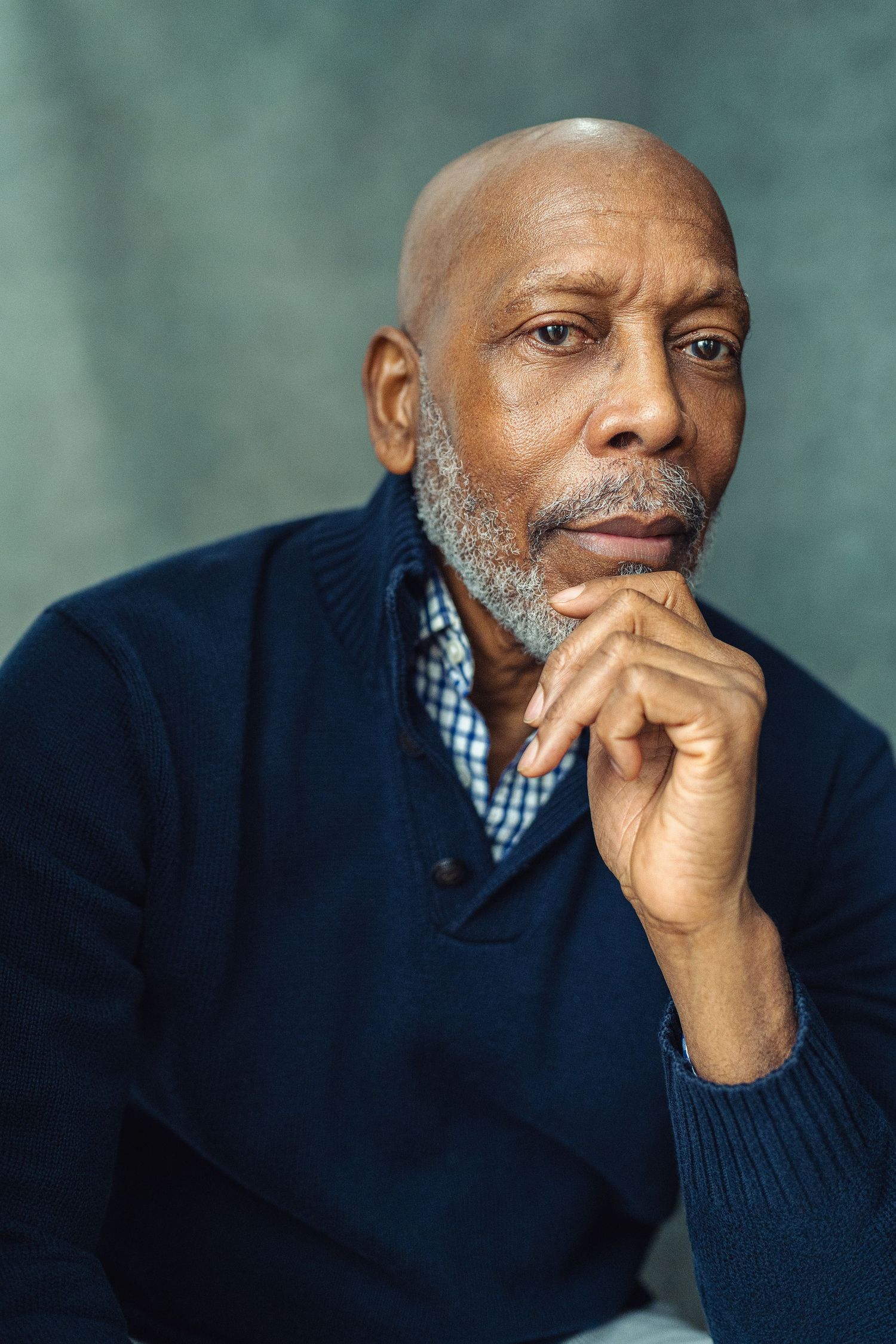 Thoughtful senior Black man with gray beard resting chin on hand, wearing navy sweater over plaid shirt, studio portrait.