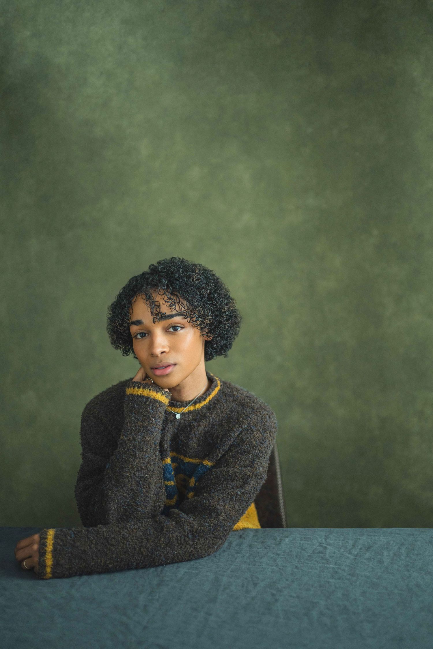 A person wearing a cozy black sweater poses against a moody green backdrop in a creative portrait series.