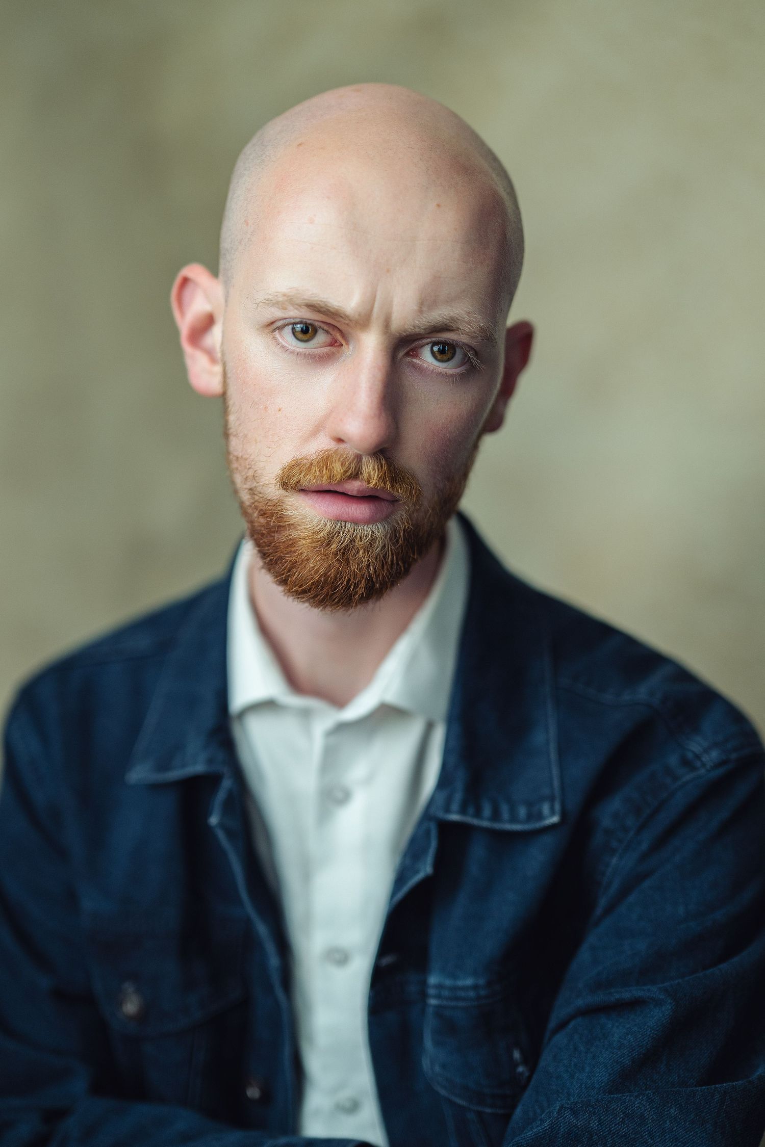 Bald man with red beard wearing denim jacket poses for portrait photo against neutral background.