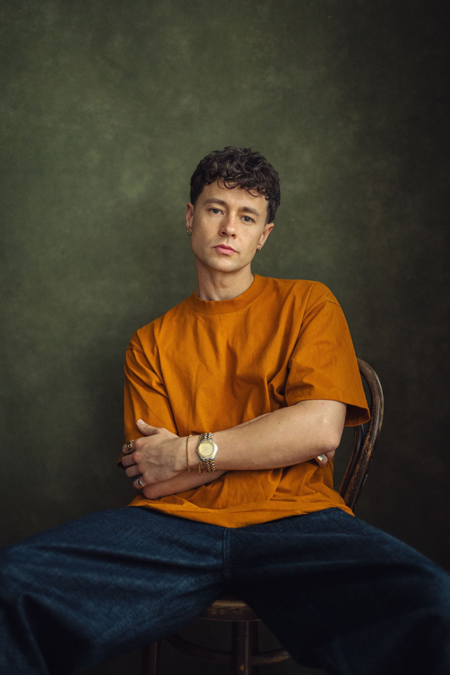 Young man in orange t-shirt sitting cross-armed on chair, wearing gold watch, against dark green studio background.