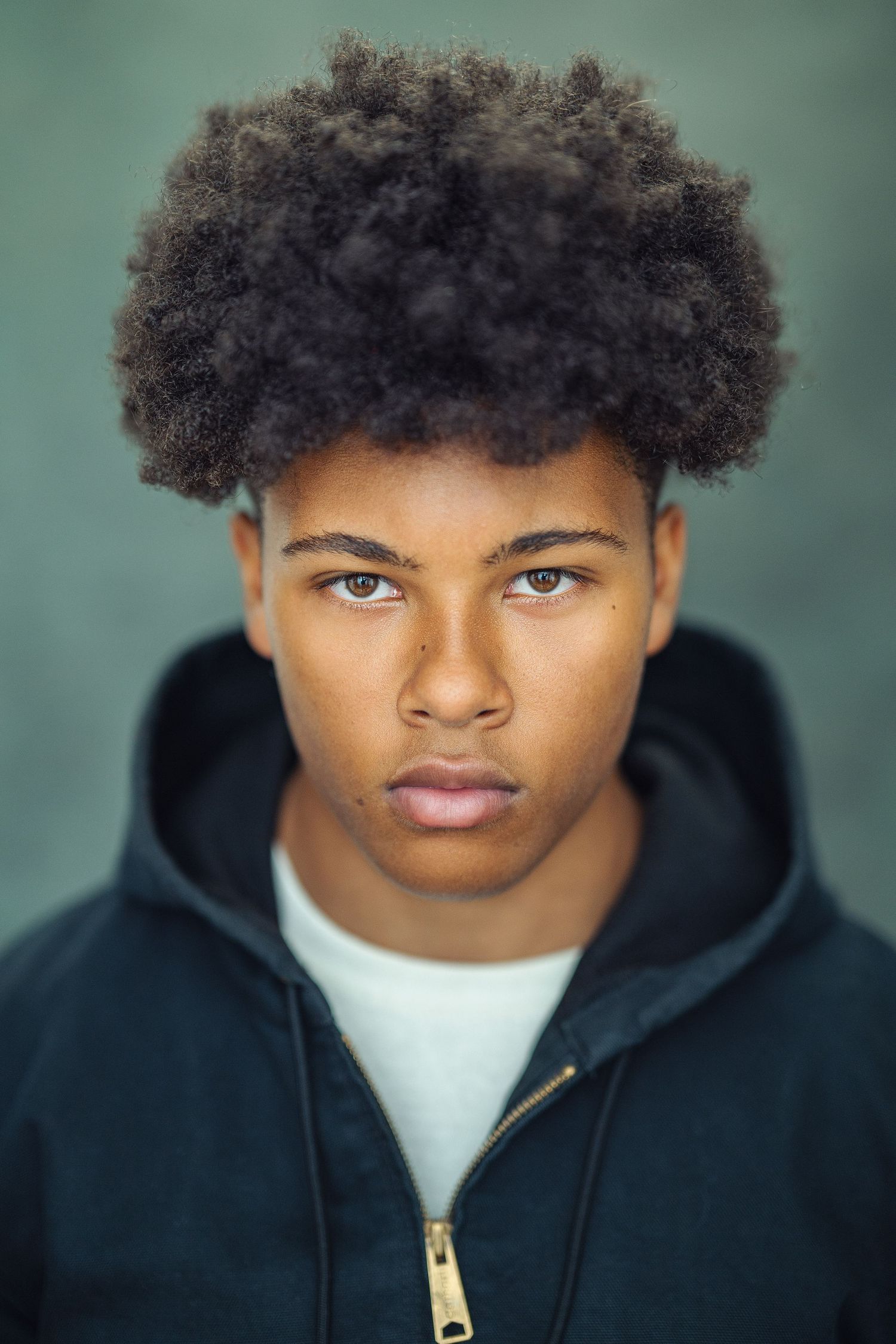 Young Black teen with natural afro hair wearing a dark hoodie, looking seriously at the camera.