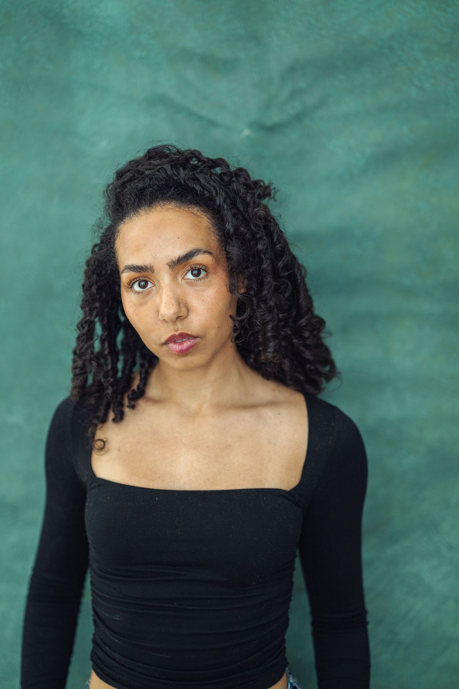 Young woman with curly hair wearing a black long-sleeve crop top, posing against a teal textured background.