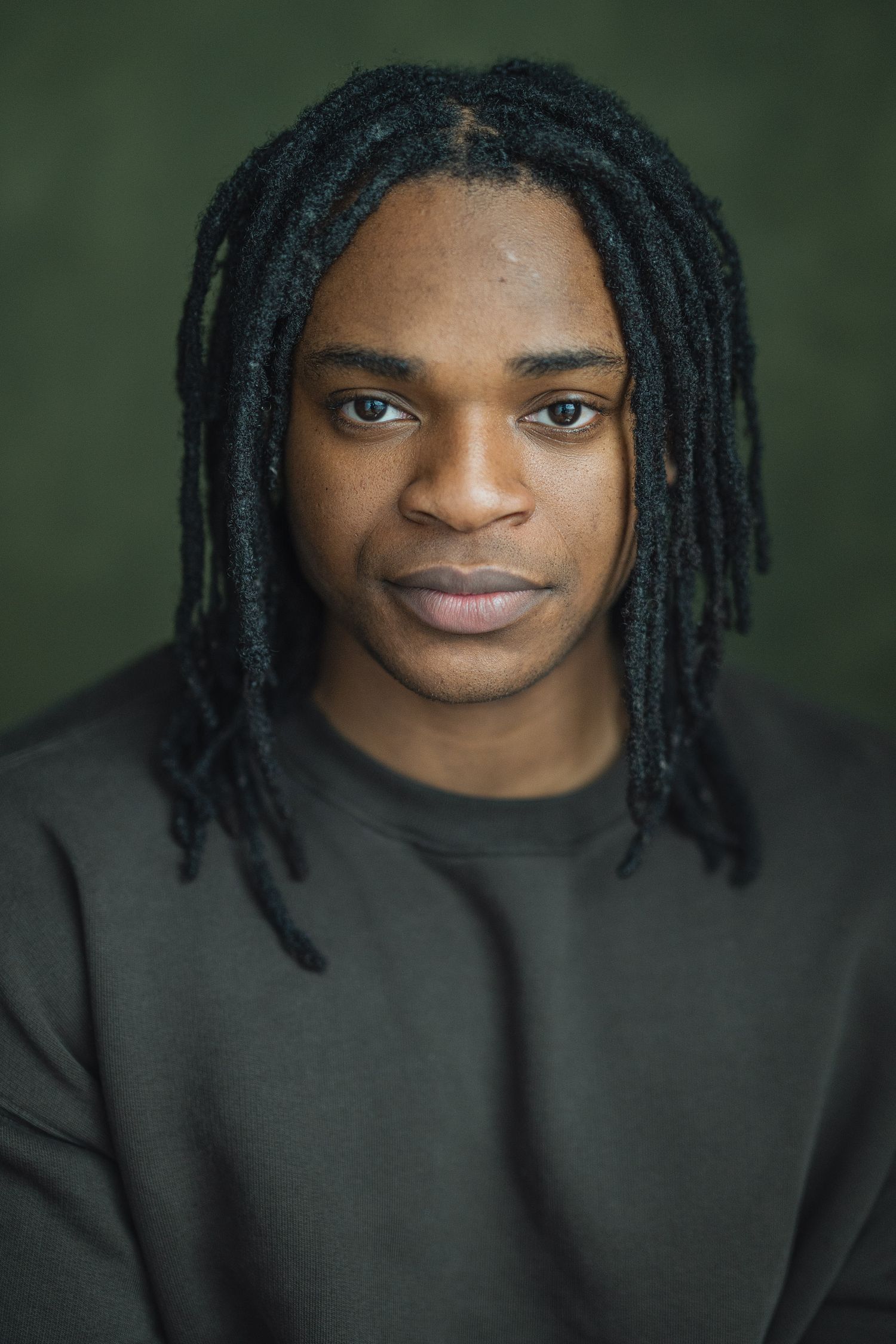 Close-up portrait of someone wearing a black sweater with dreadlocks against a dark green background.