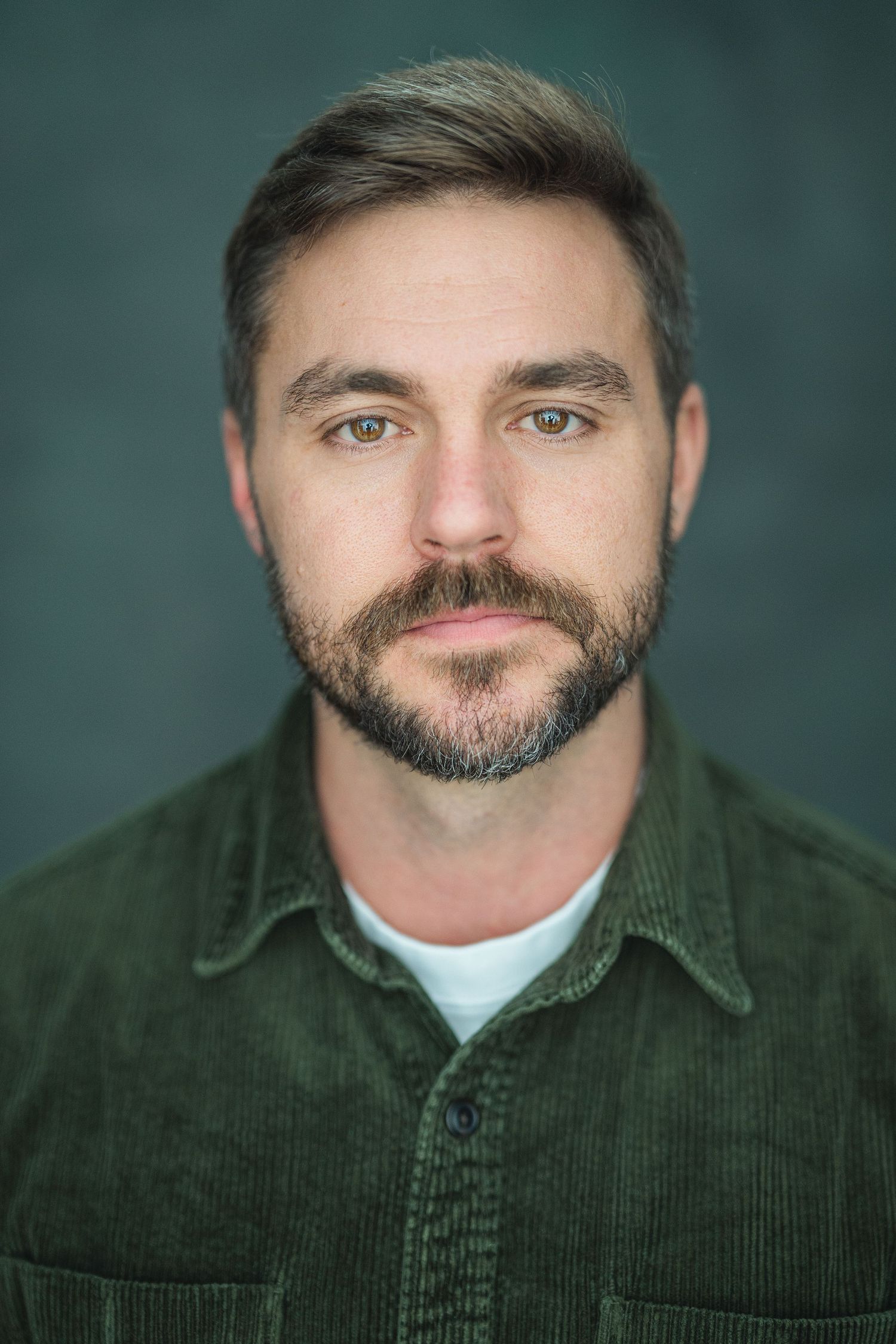 Bearded man with short brown hair wearing a green corduroy shirt, posing against a dark teal background.