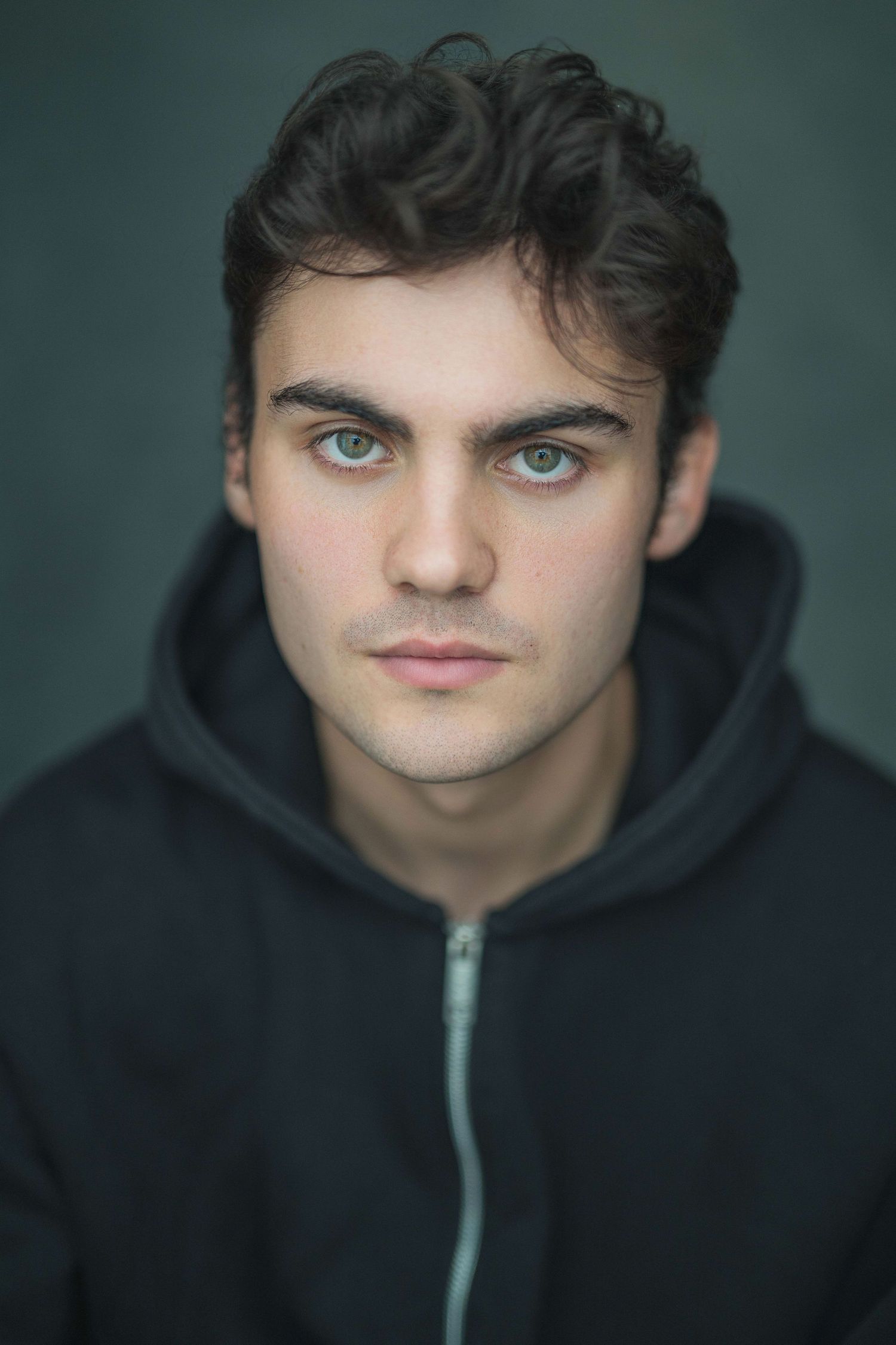Portrait of someone wearing a black hoodie with striking blue eyes against a dark green background.