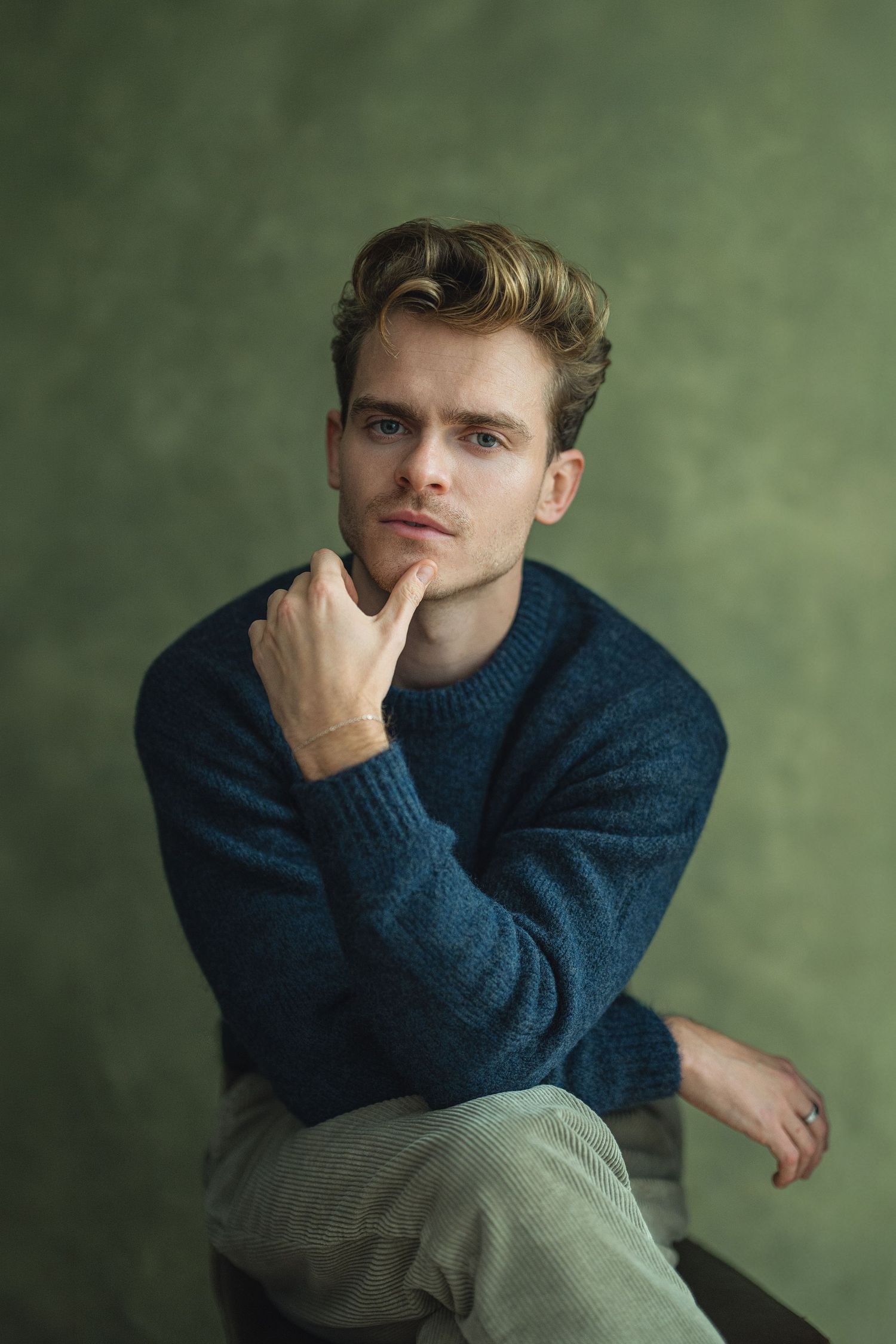 A person in a navy blue sweater sits thoughtfully with hand on chin against a green background.