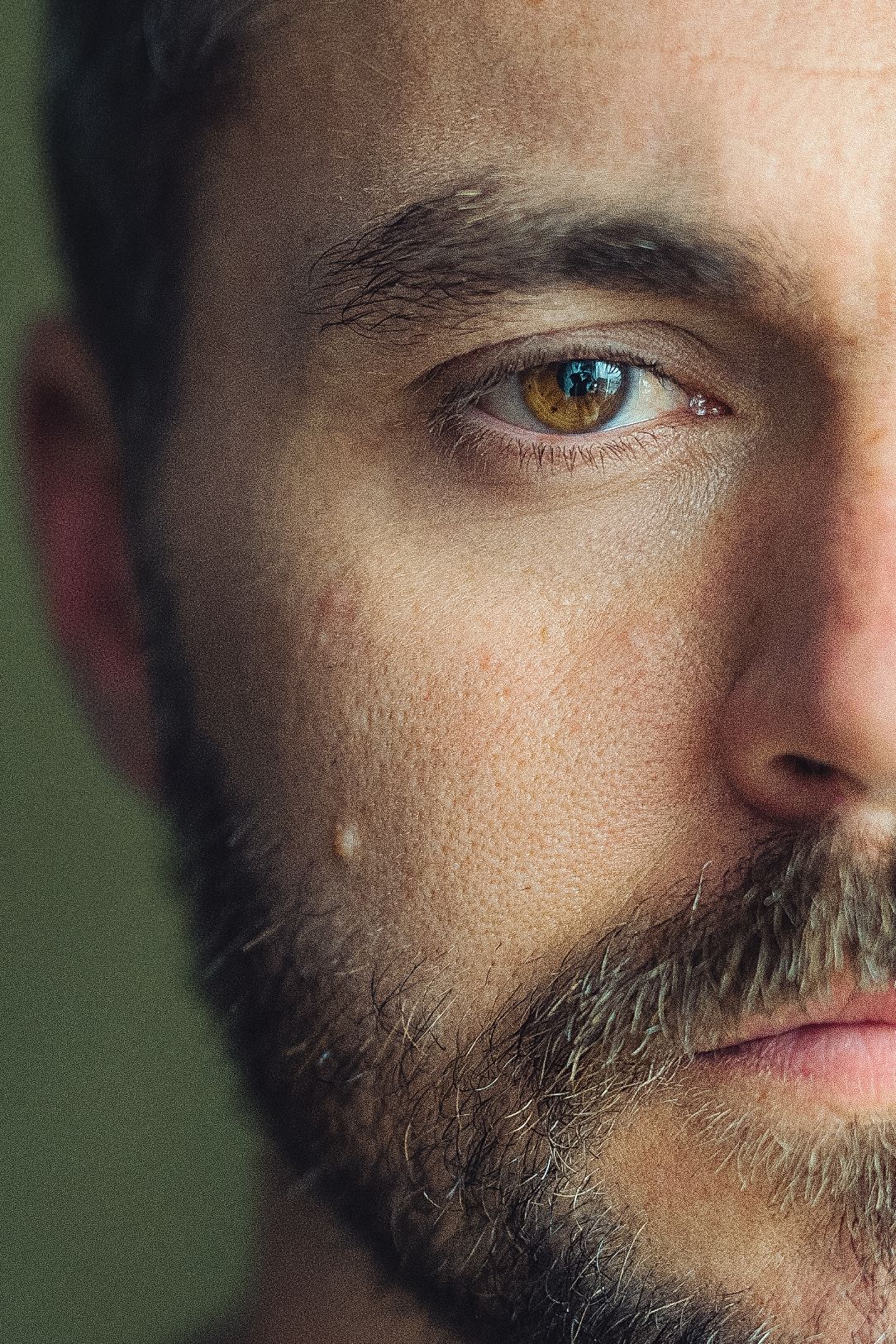 Close-up portrait of a bearded man with hazel eyes on a dark green background.