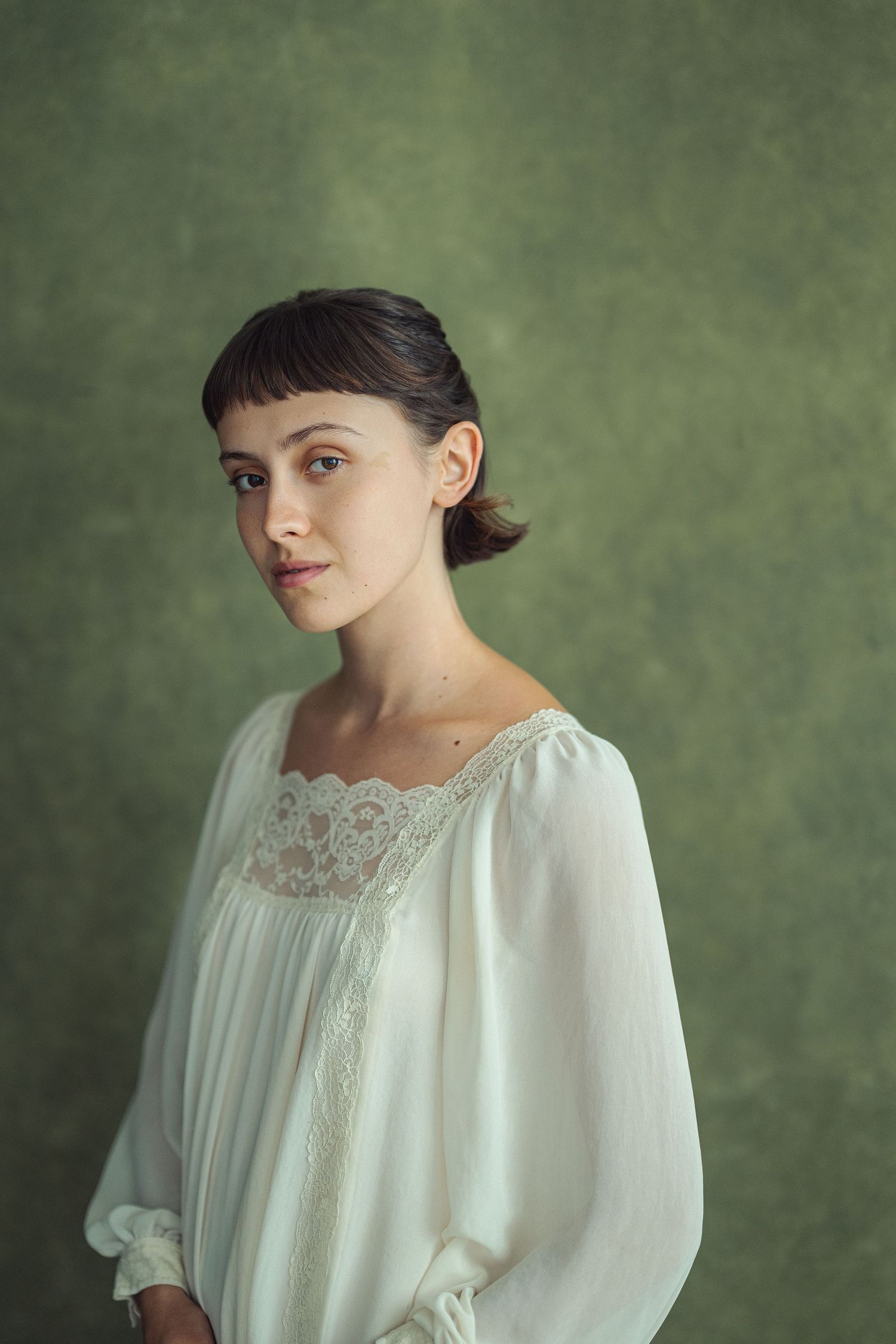 Young woman in white lace blouse poses against green backdrop in elegant portrait photography.