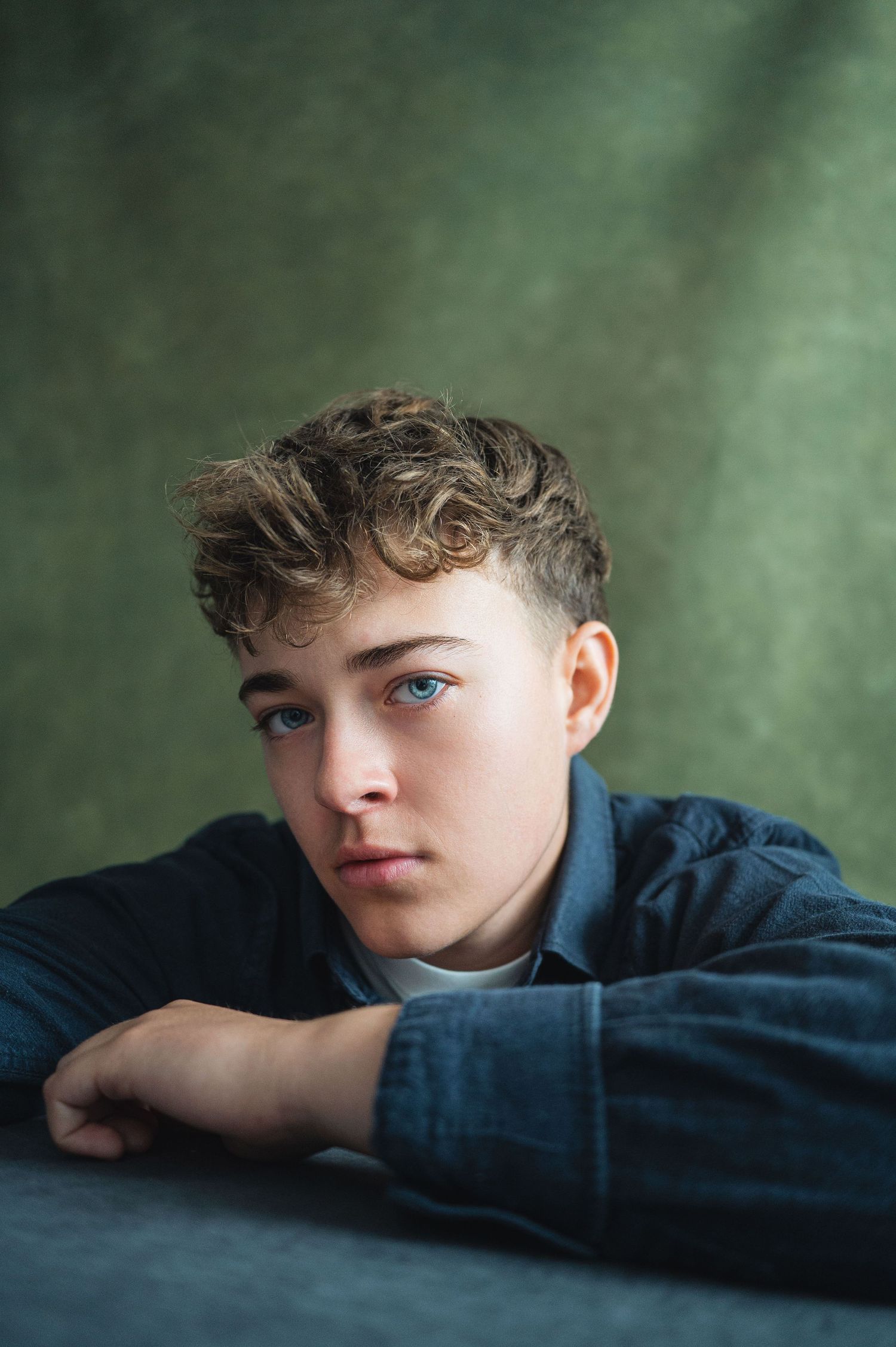 A young teen boy with curly hair and blue eyes rests his arms on a surface, posing for a portrait against a green backdrop.
