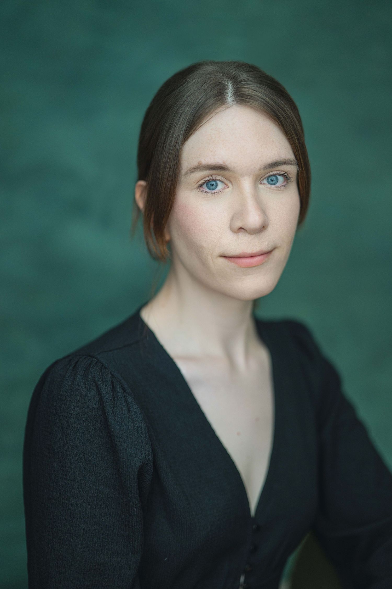 Young woman with blue eyes and brown hair posing against a dark green background.