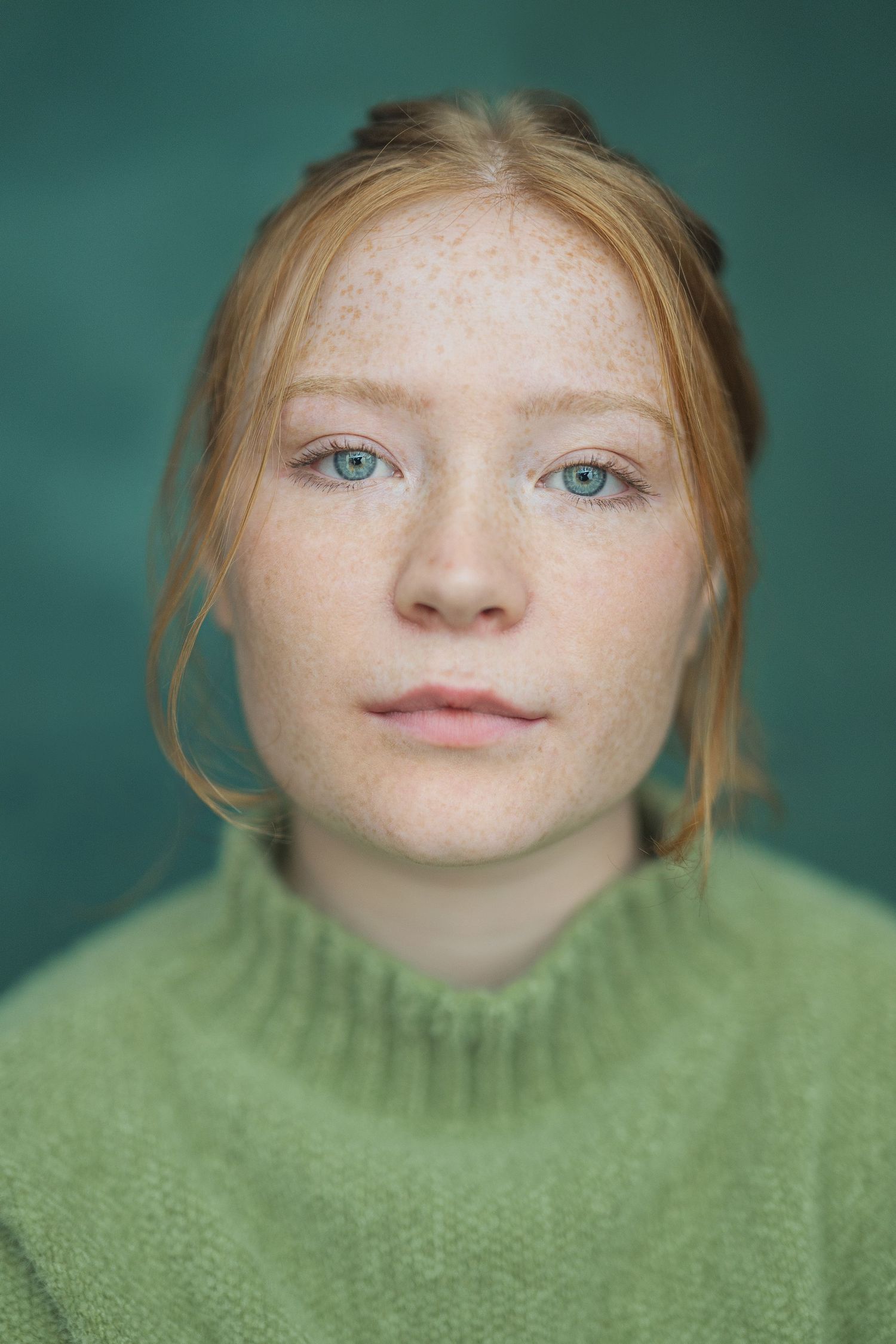 Close-up portrait showing bright blue eyes and freckles against teal background while wearing a green knit sweater.