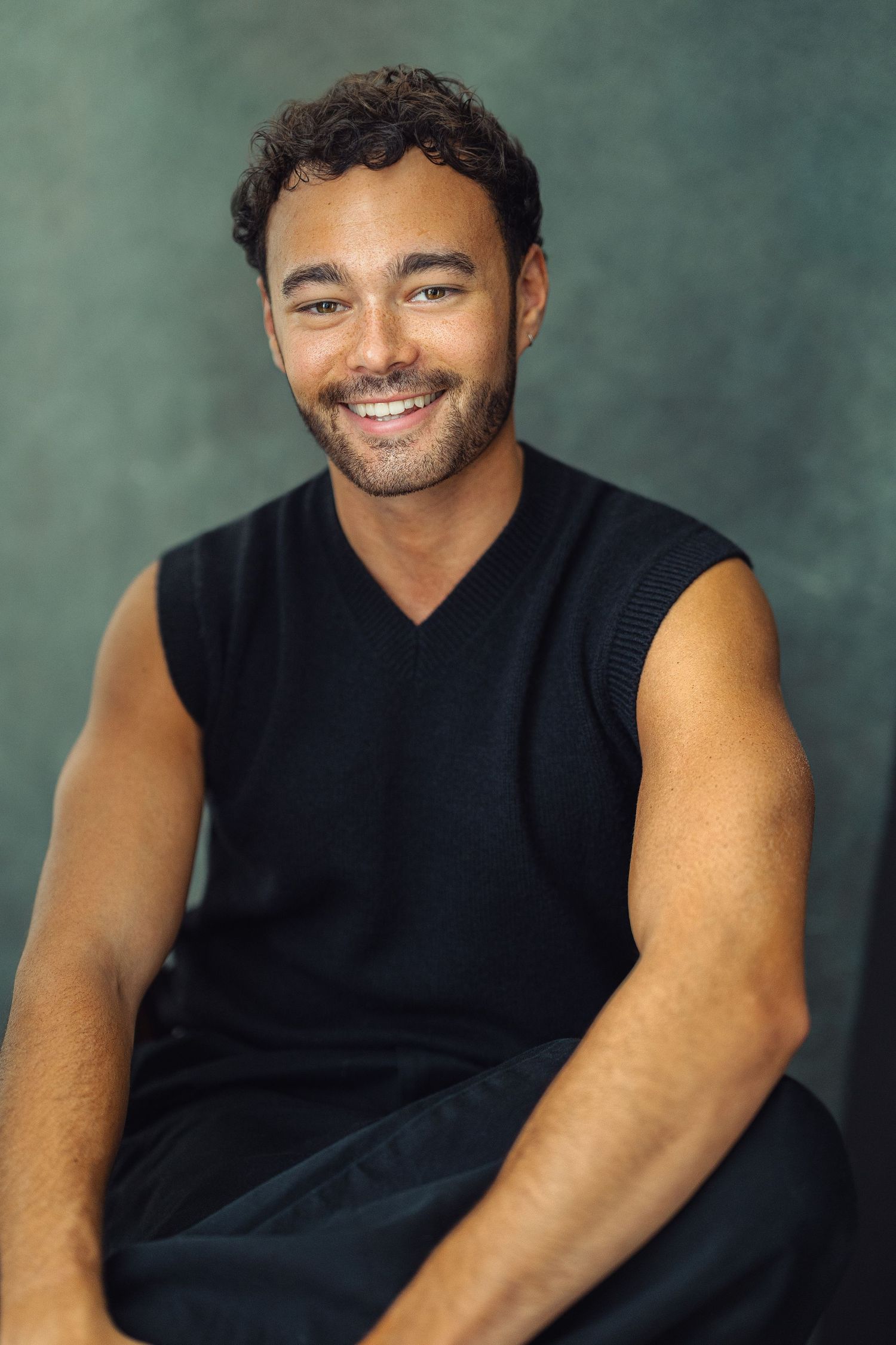Smiling young man with curly hair wearing a black sleeveless top, seated against a dark green background.
