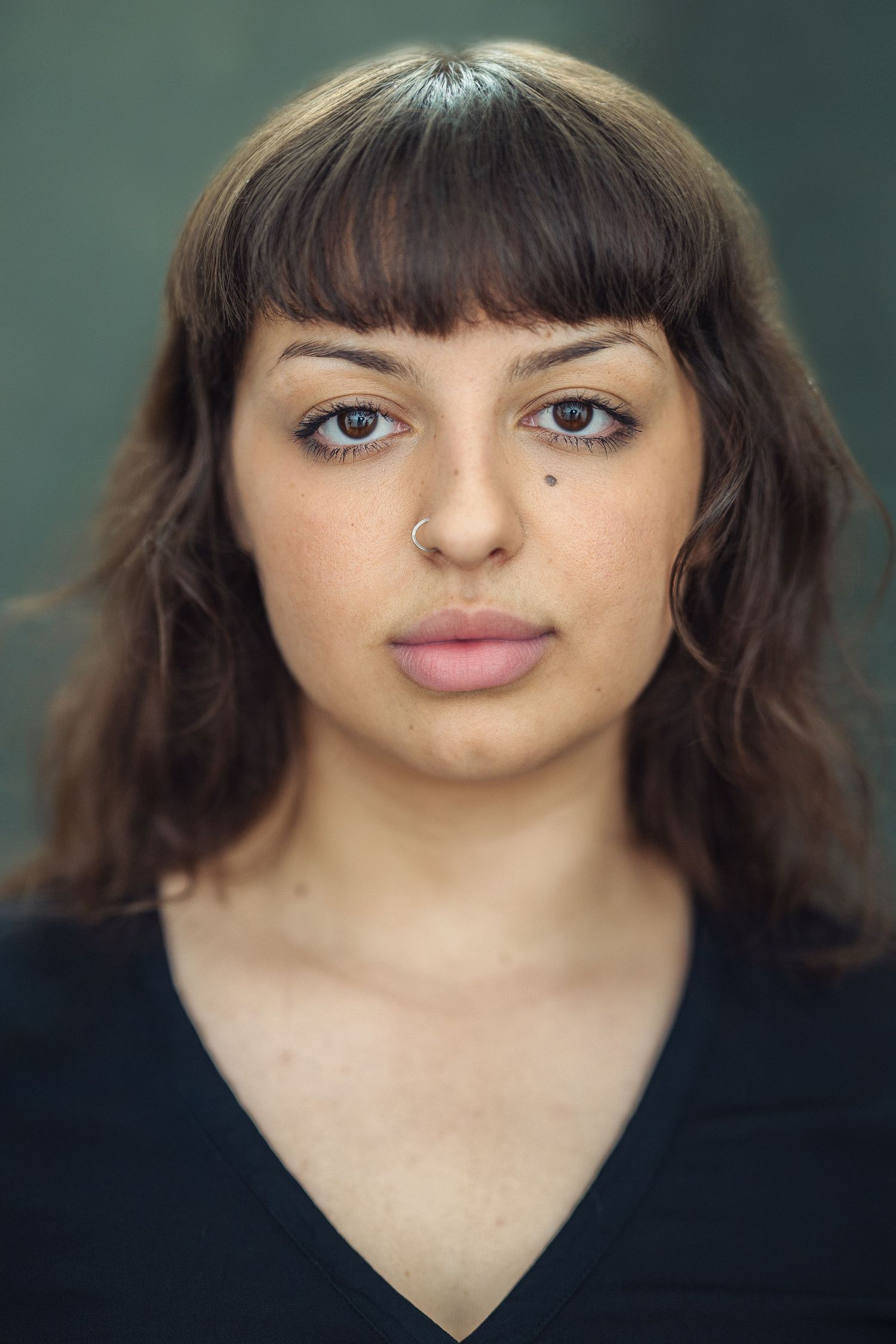 Young woman with brown wavy hair and bangs, wearing a black top, posing for a portrait against a dark background.