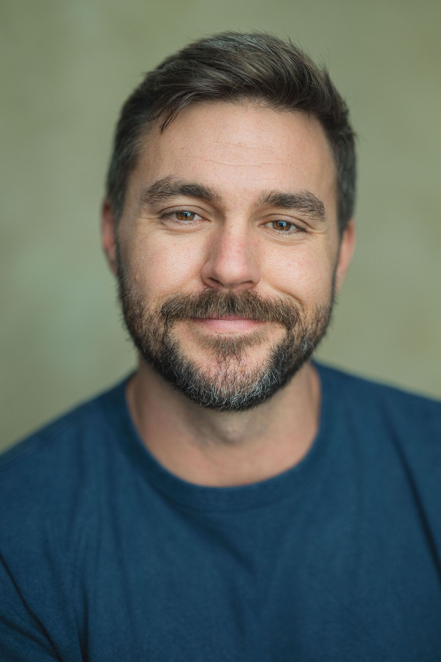 Smiling bearded man with short brown hair wearing a blue t-shirt against a green background.