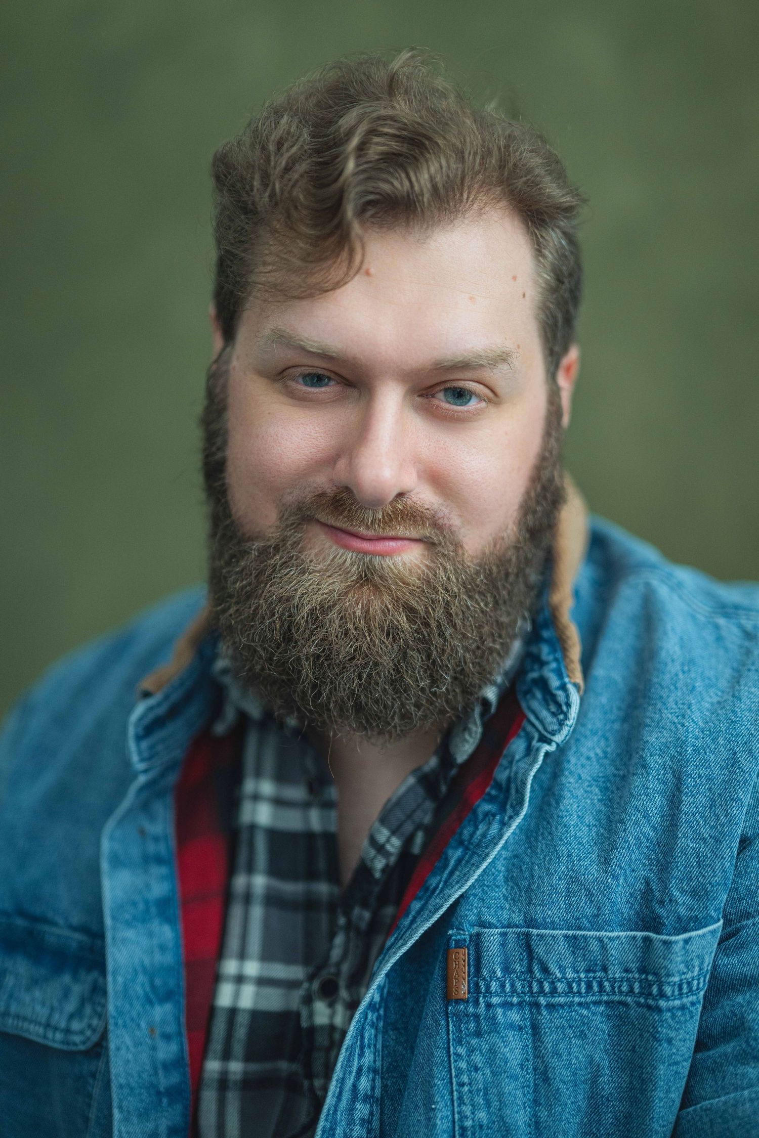 A person wearing a denim jacket and plaid shirt smiles warmly against a blurred green background.