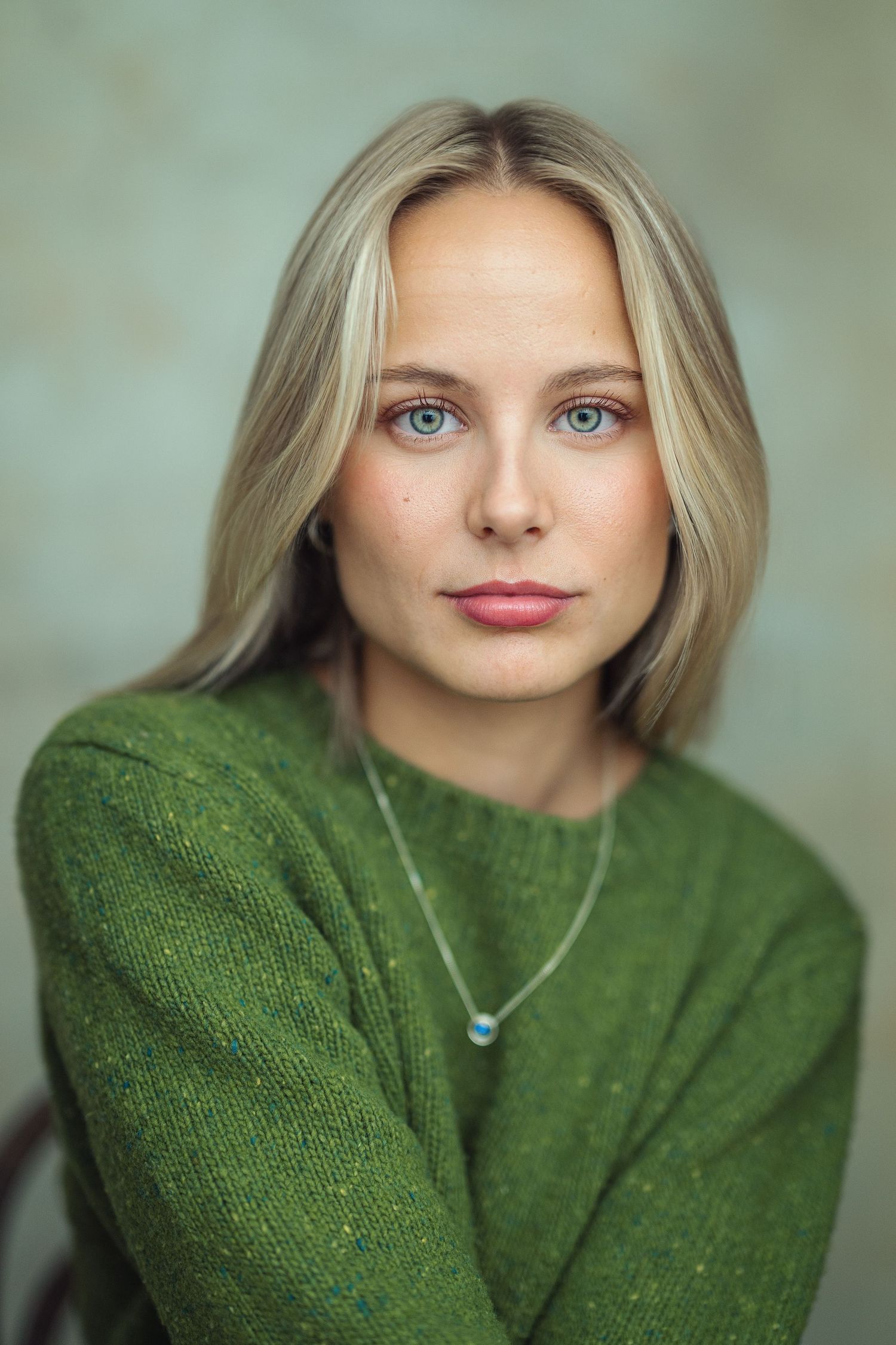 Young blonde woman with blue eyes wearing a green knit sweater and delicate necklace poses for a portrait photo.