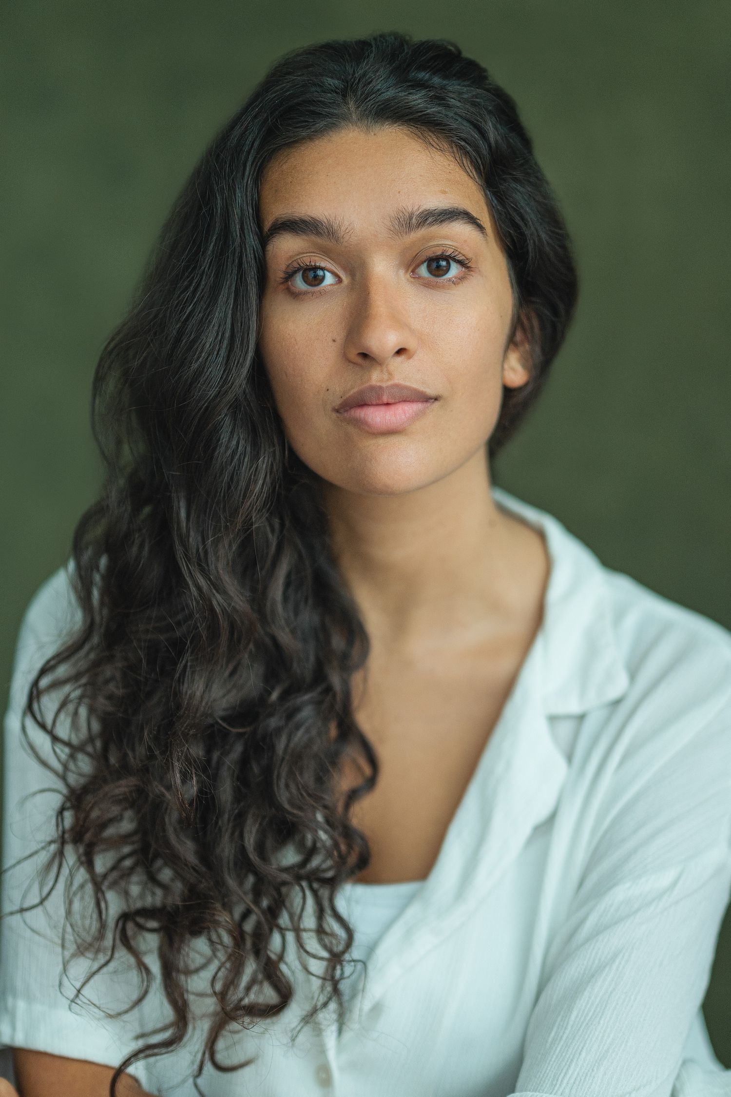 Portrait of someone with long dark curly hair wearing a white top against a green background.