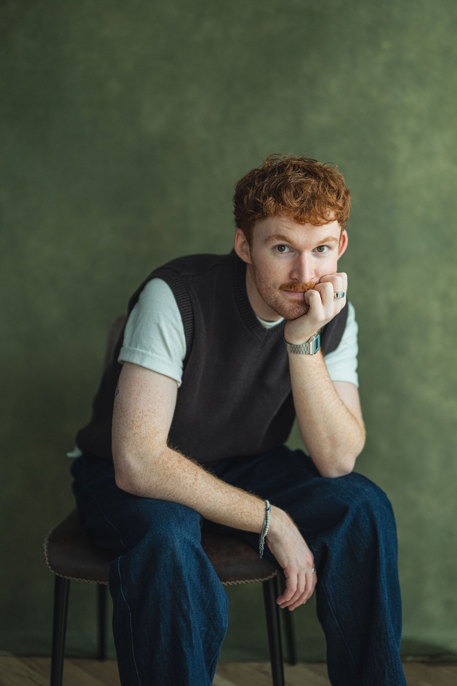 A person with red hair sits pensively in a chair against a green wall wearing dark clothing and a light shirt.