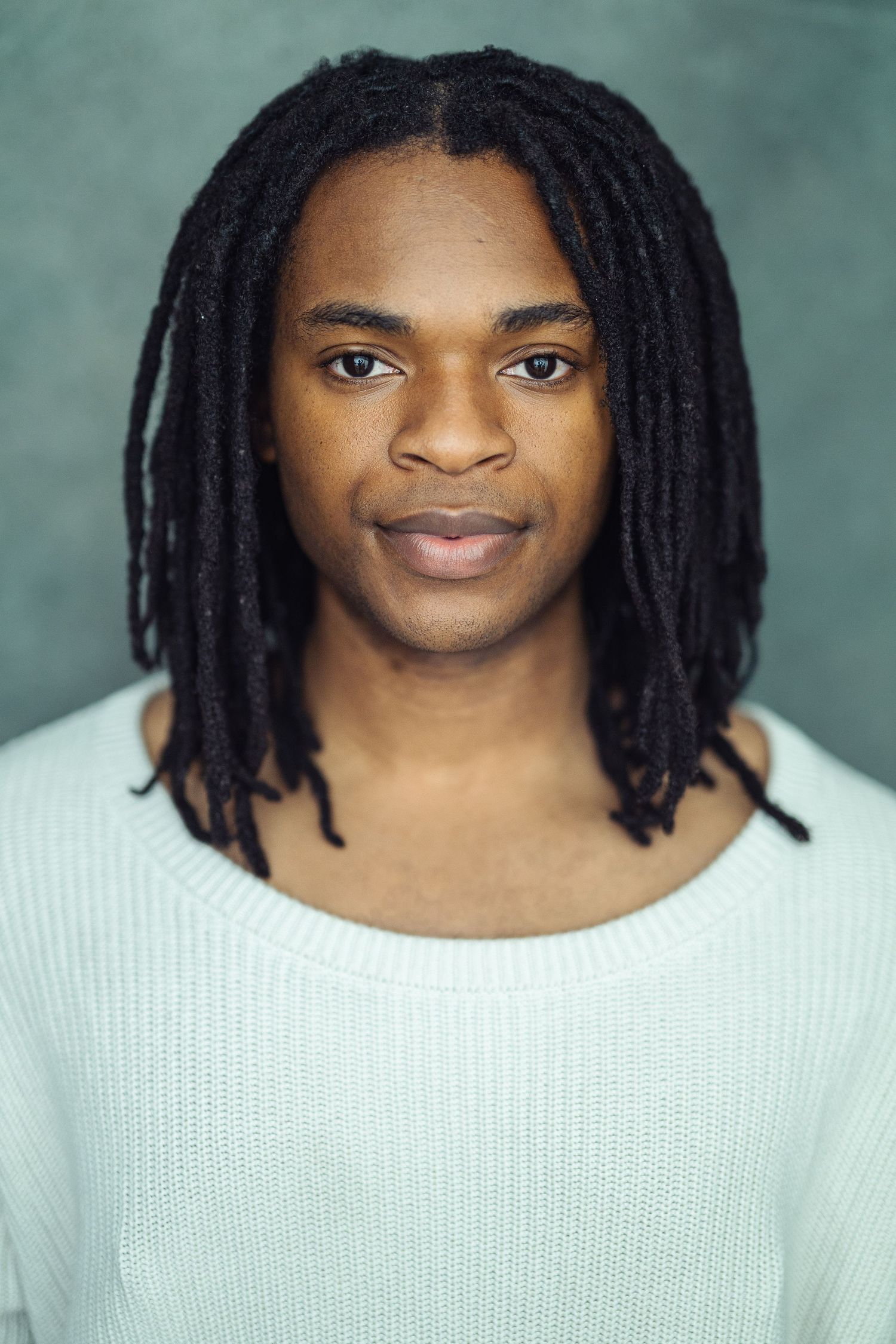 Young Black man with long dreadlocks wearing a light blue sweater, posing for a professional headshot photo.