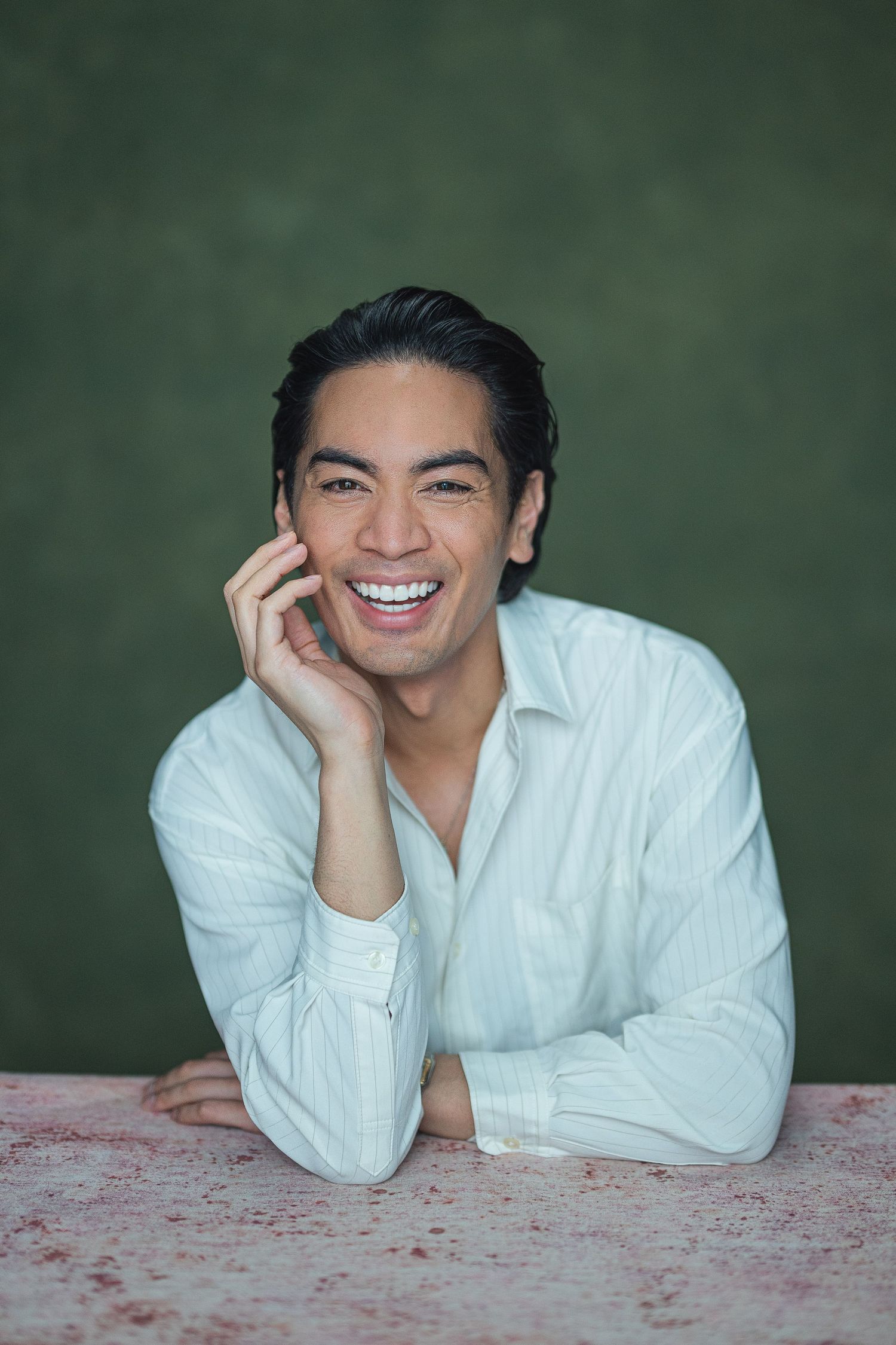 Smiling young Asian man in white striped shirt leaning on table against green background.