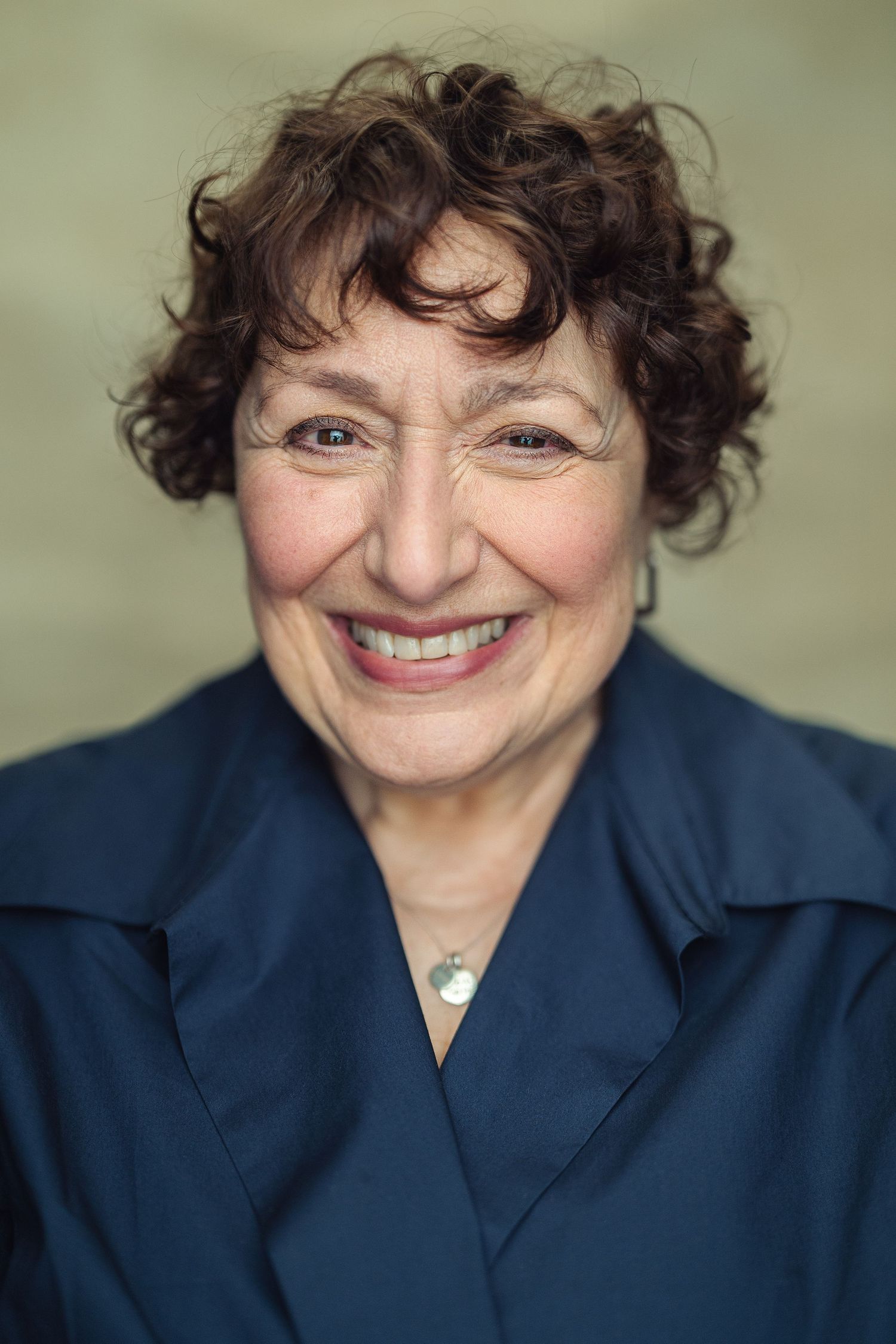 Smiling older woman with curly brown hair wearing a navy blue blouse and silver necklace, professional headshot photo.