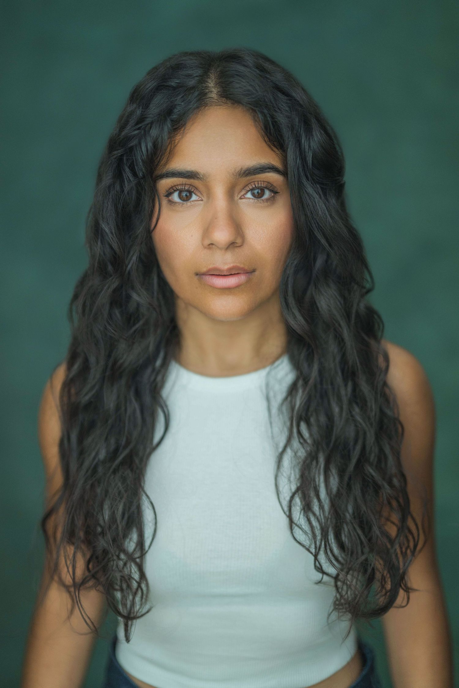 Person with long dark wavy hair wearing a white sleeveless top poses against a teal colored background.