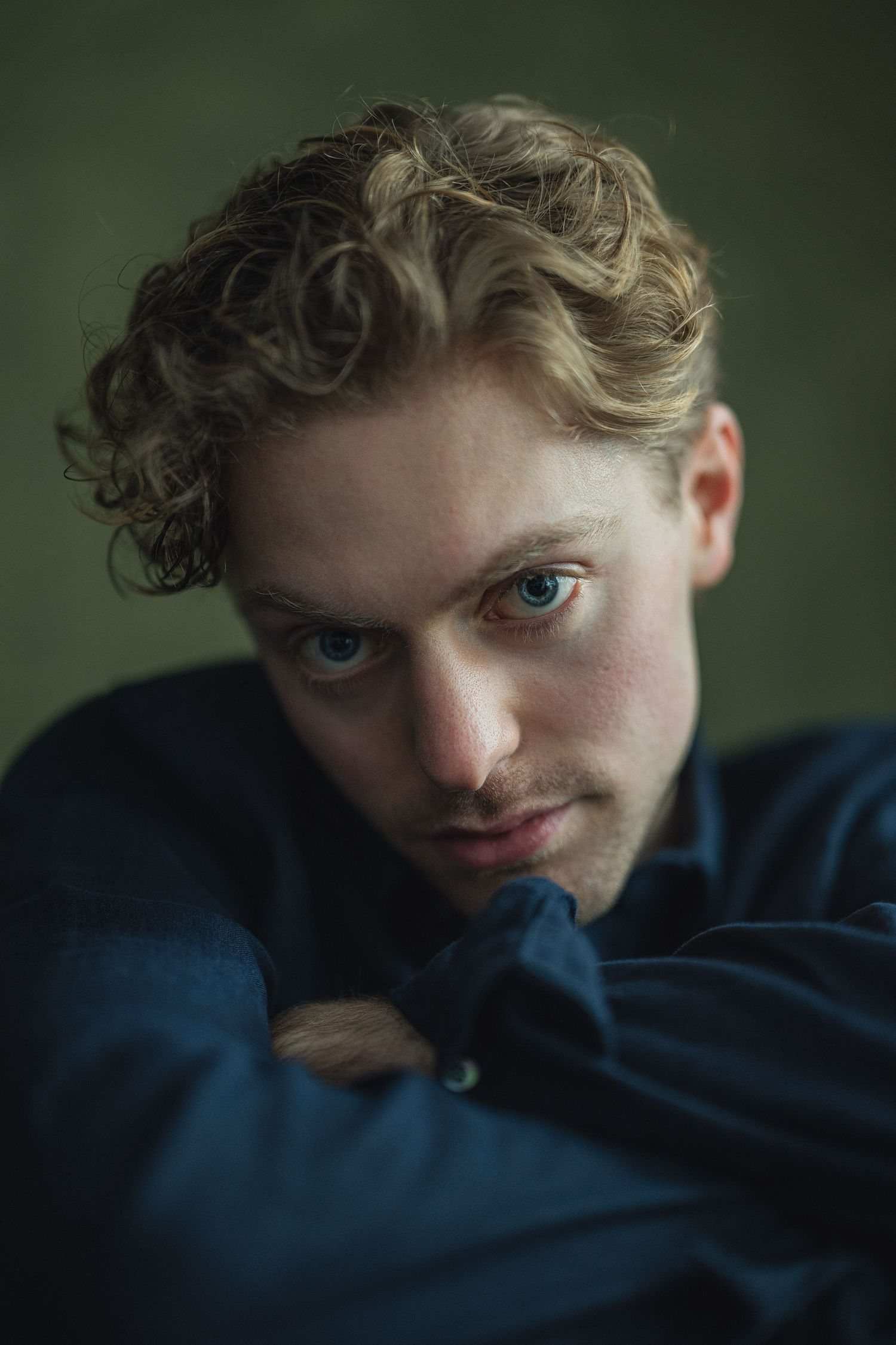 Dramatic portrait with curly blonde hair and blue sweater against dark green background in moody lighting.
