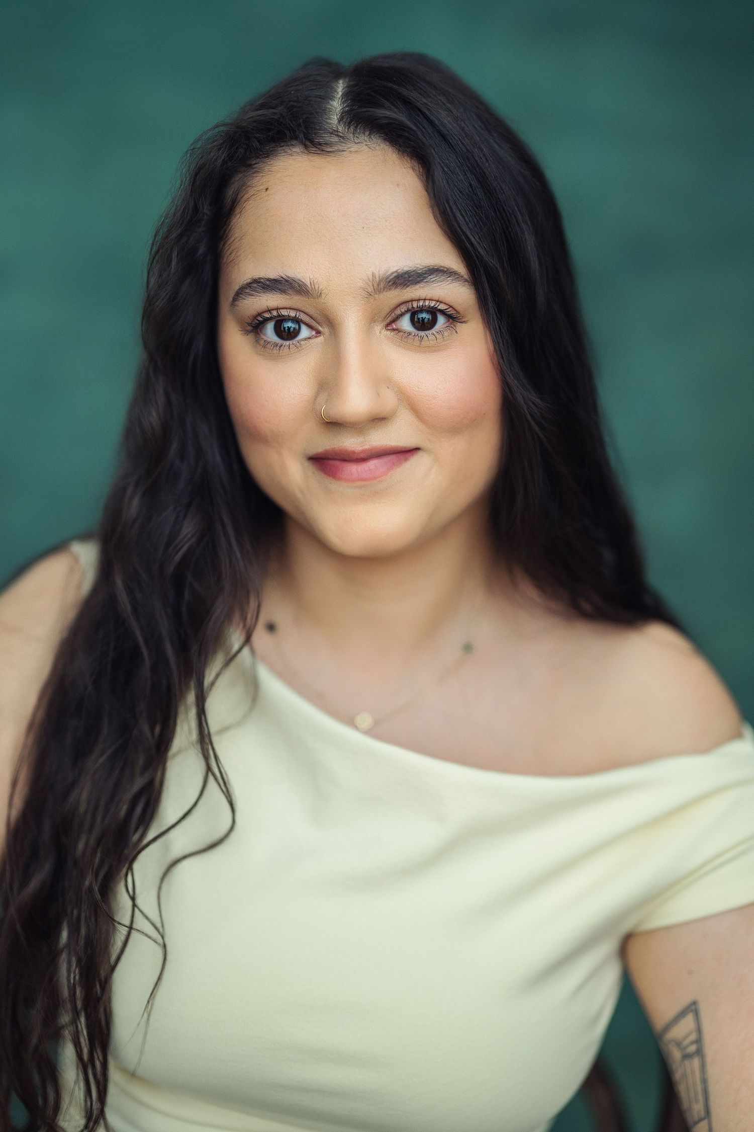 Young woman with long dark hair in cream off-shoulder top smiling against teal background.