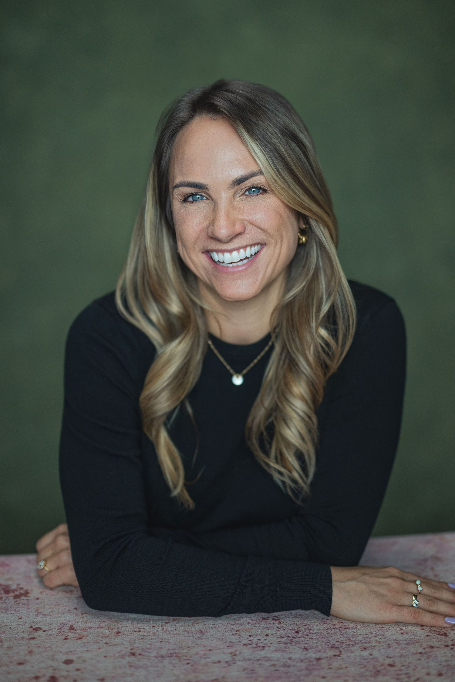 Smiling woman with blonde hair wearing a black top and green necklace, posing against a green background.