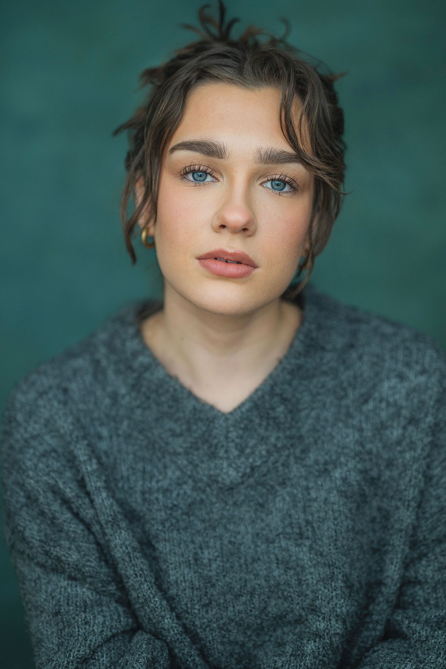 Portrait against teal background showing striking blue eyes and dark gray sweater in soft natural lighting.