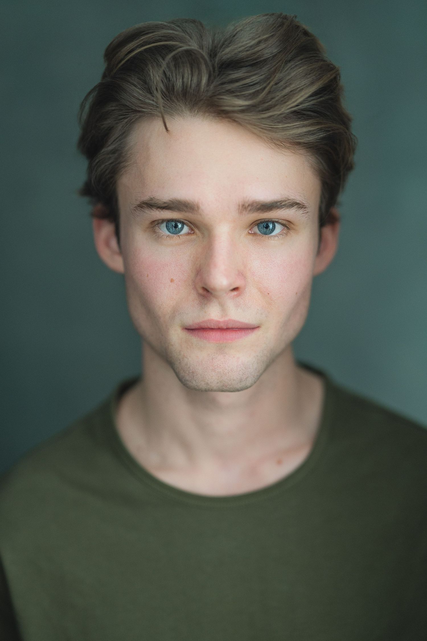 Professional headshot portrait against teal background showing person in olive green shirt with styled hair and blue eyes.