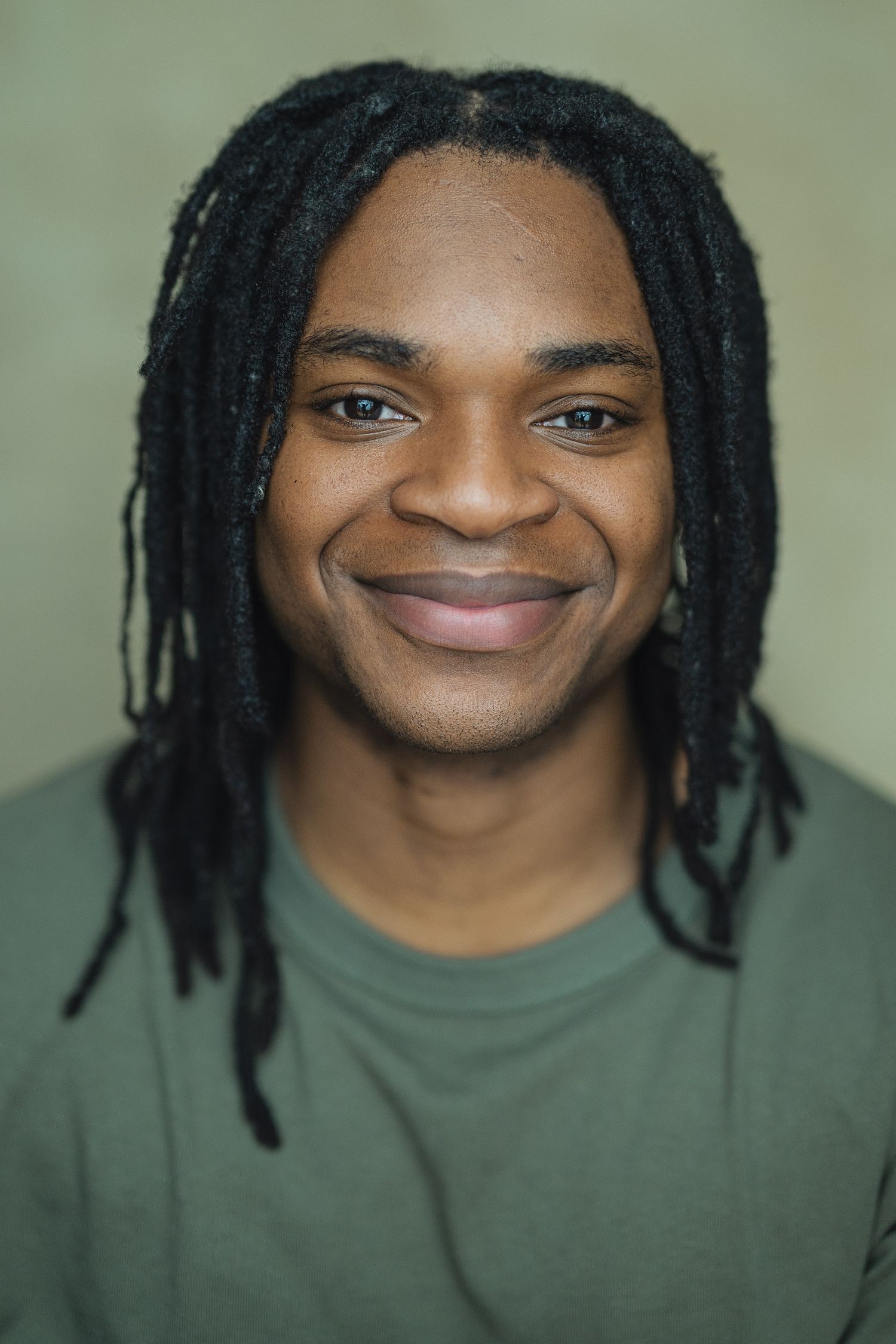 Portrait of a person with dreadlocks wearing a green shirt smiling warmly against a neutral background.