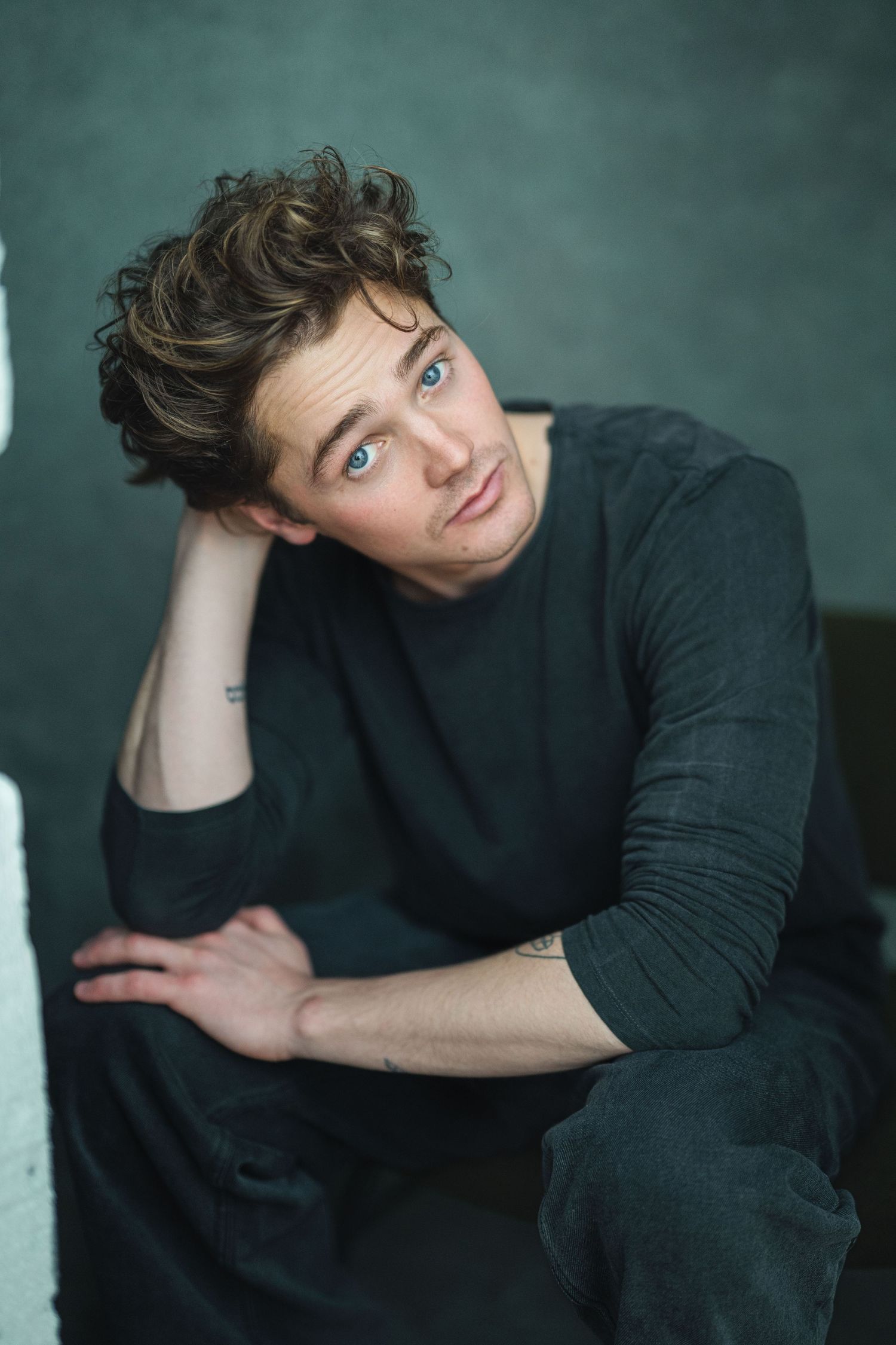 A person in a black sweater poses against a teal background in a moody portrait series.