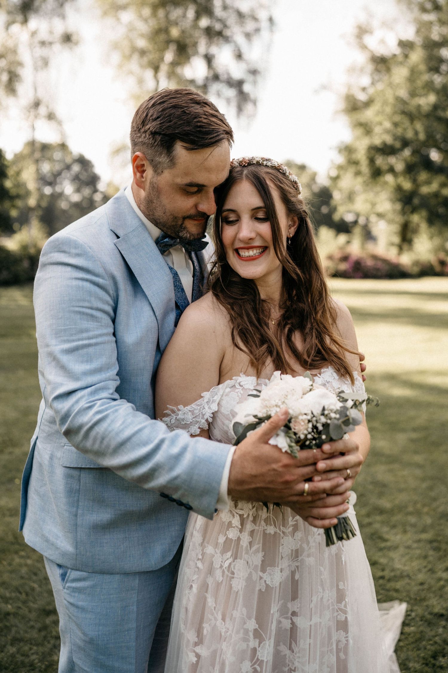 A romantic outdoor wedding moment with couple embracing in a sunny garden setting, bride in lace gown holding white flowers.
