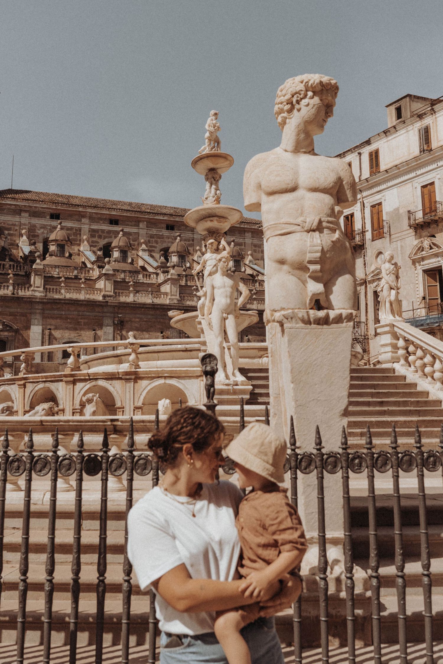 European plaza with ornate baroque statues and fountains featuring classical stone sculptures and architecture in warm tones.