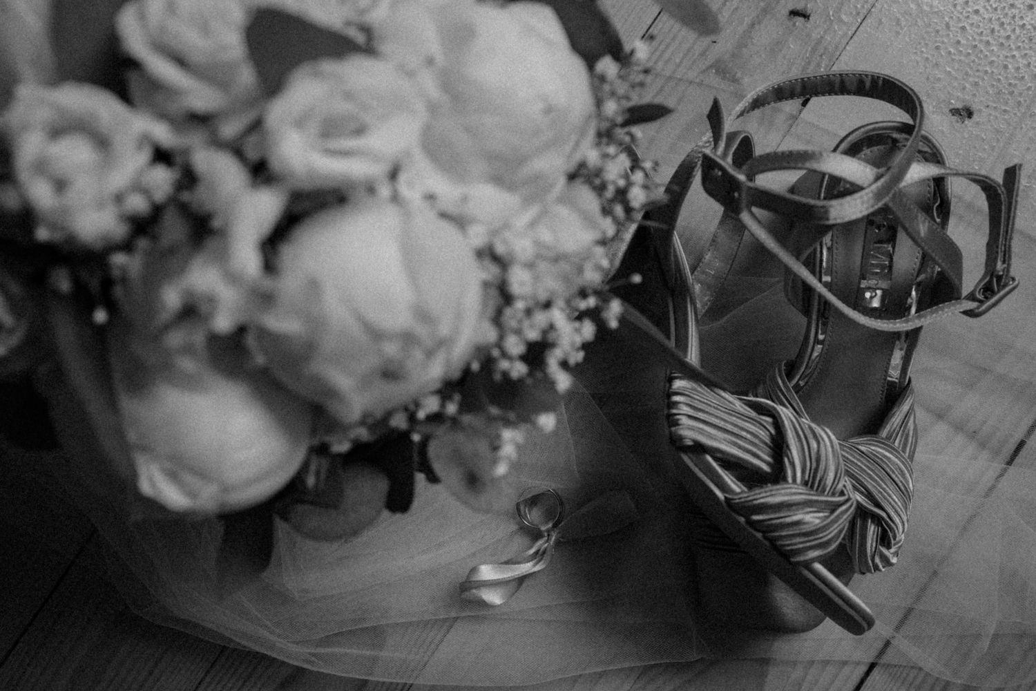 A black and white photograph of roses in a vase next to a pair of dress shoes on a wooden floor.