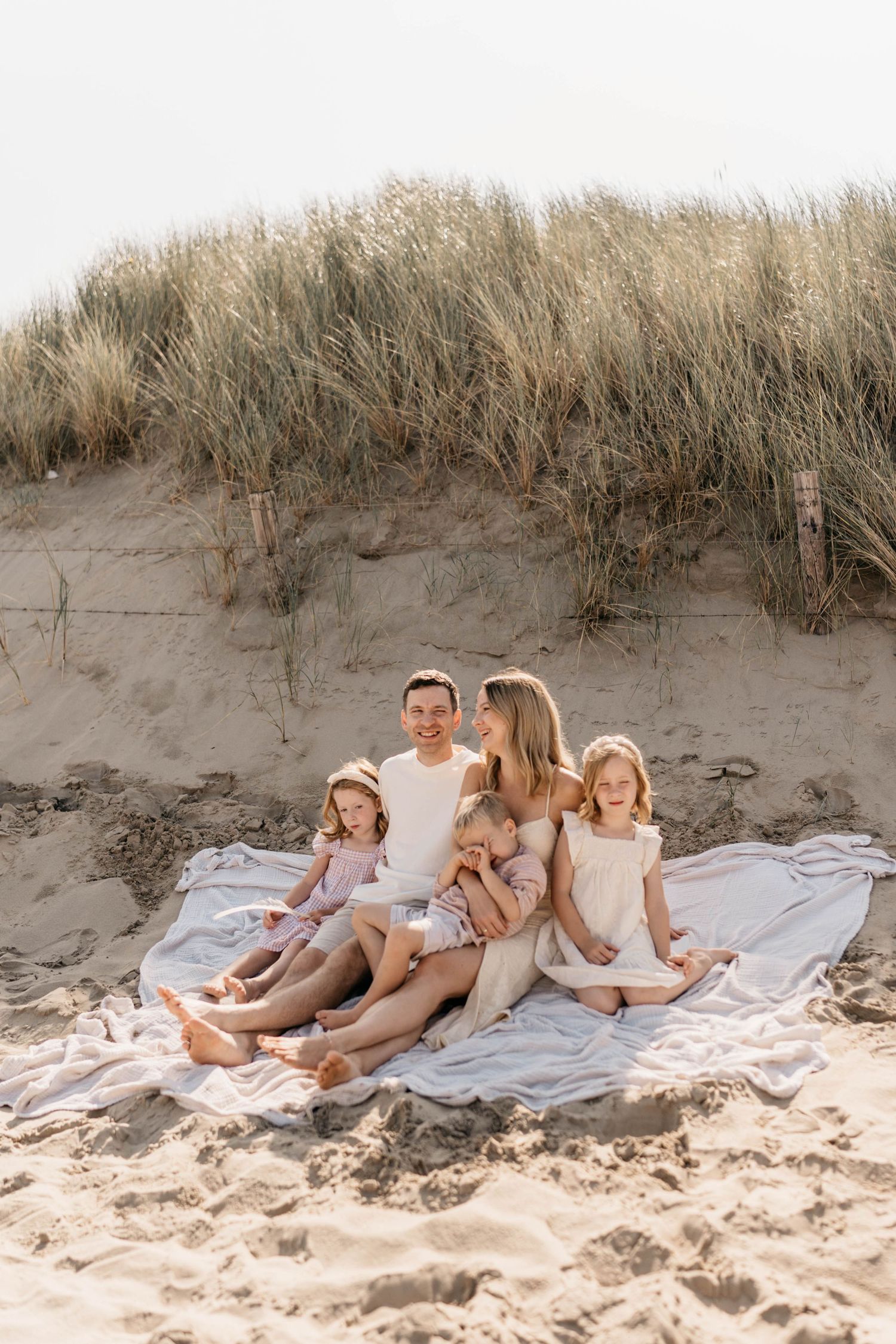 A family sits together on a white blanket at the beach with tall grass-covered dunes in the background.