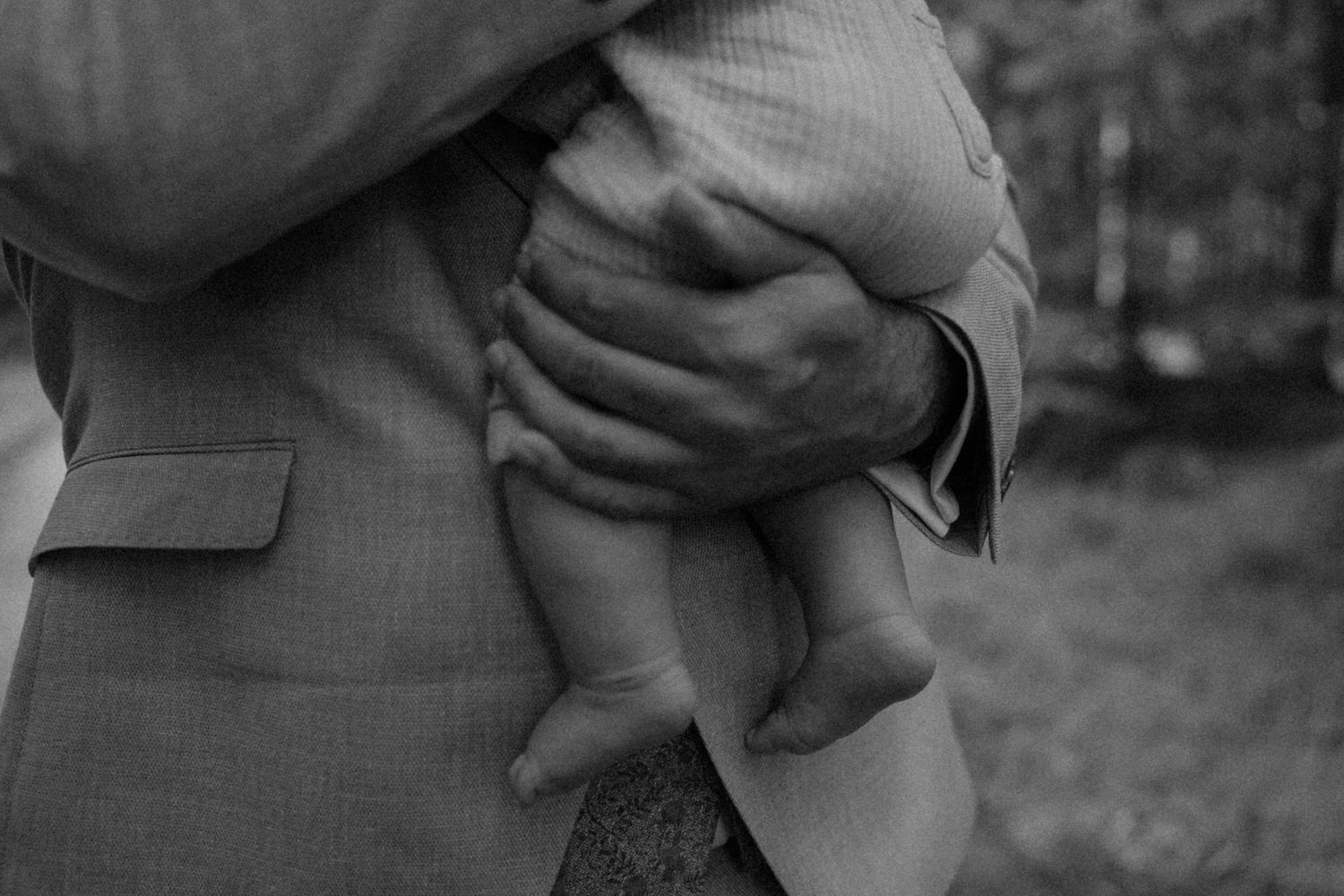 A black and white photo of a couple embracing, showing the details of formal attire against a natural outdoor setting.