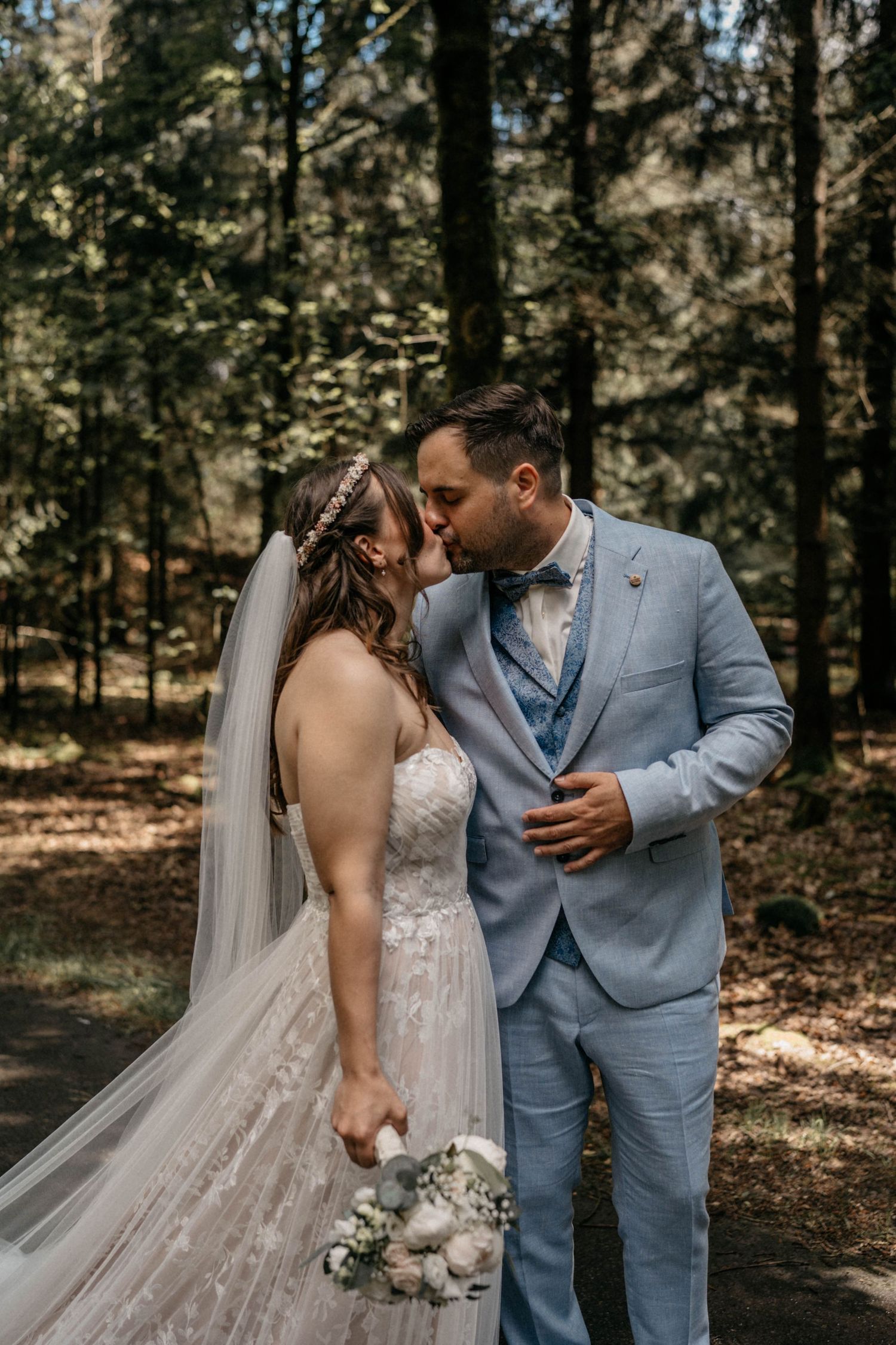 Newlywed couple sharing an intimate moment in a sunlit forest wearing light blue suit and white wedding dress.