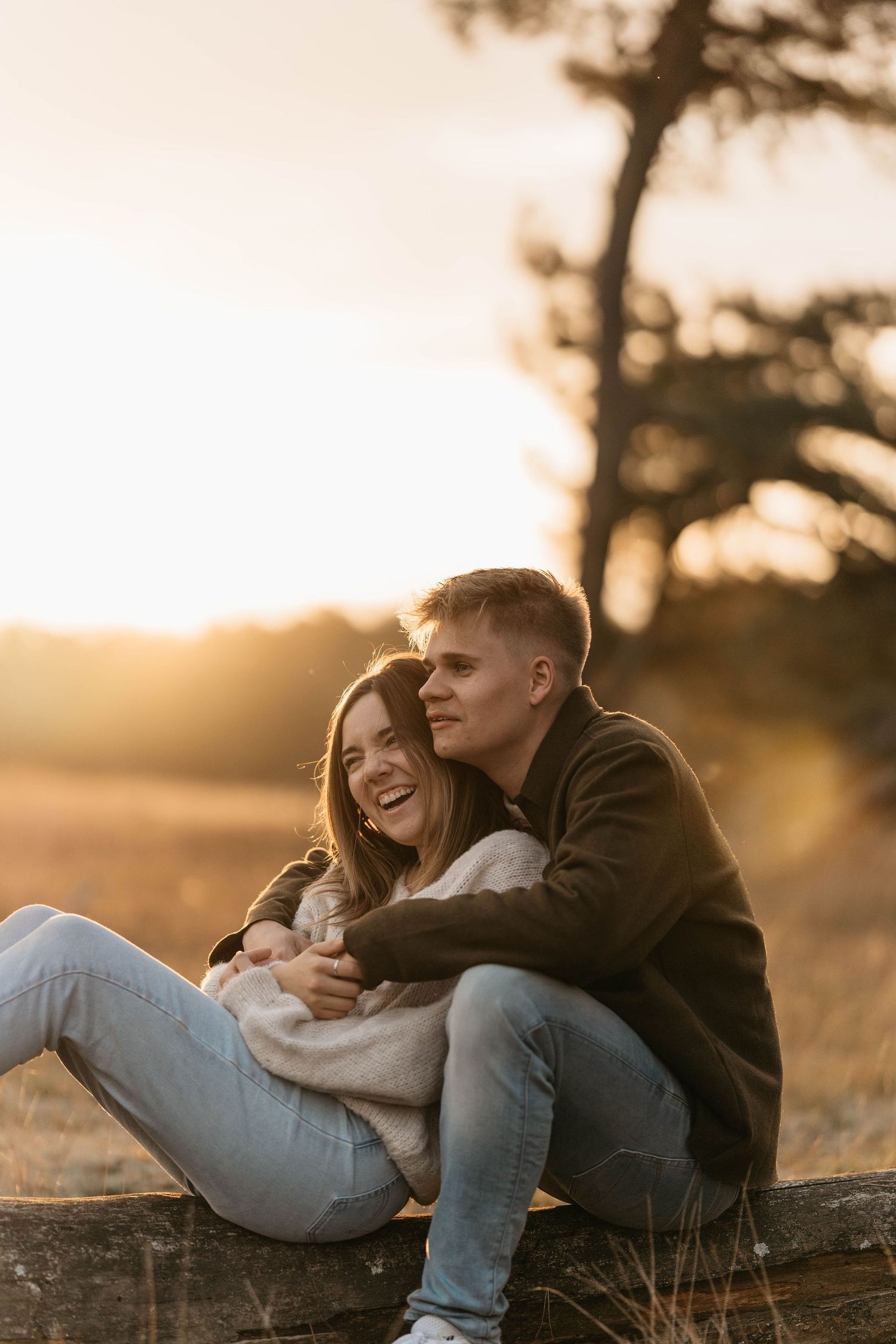 Couple laughing together while sitting on wooden fence at sunset.