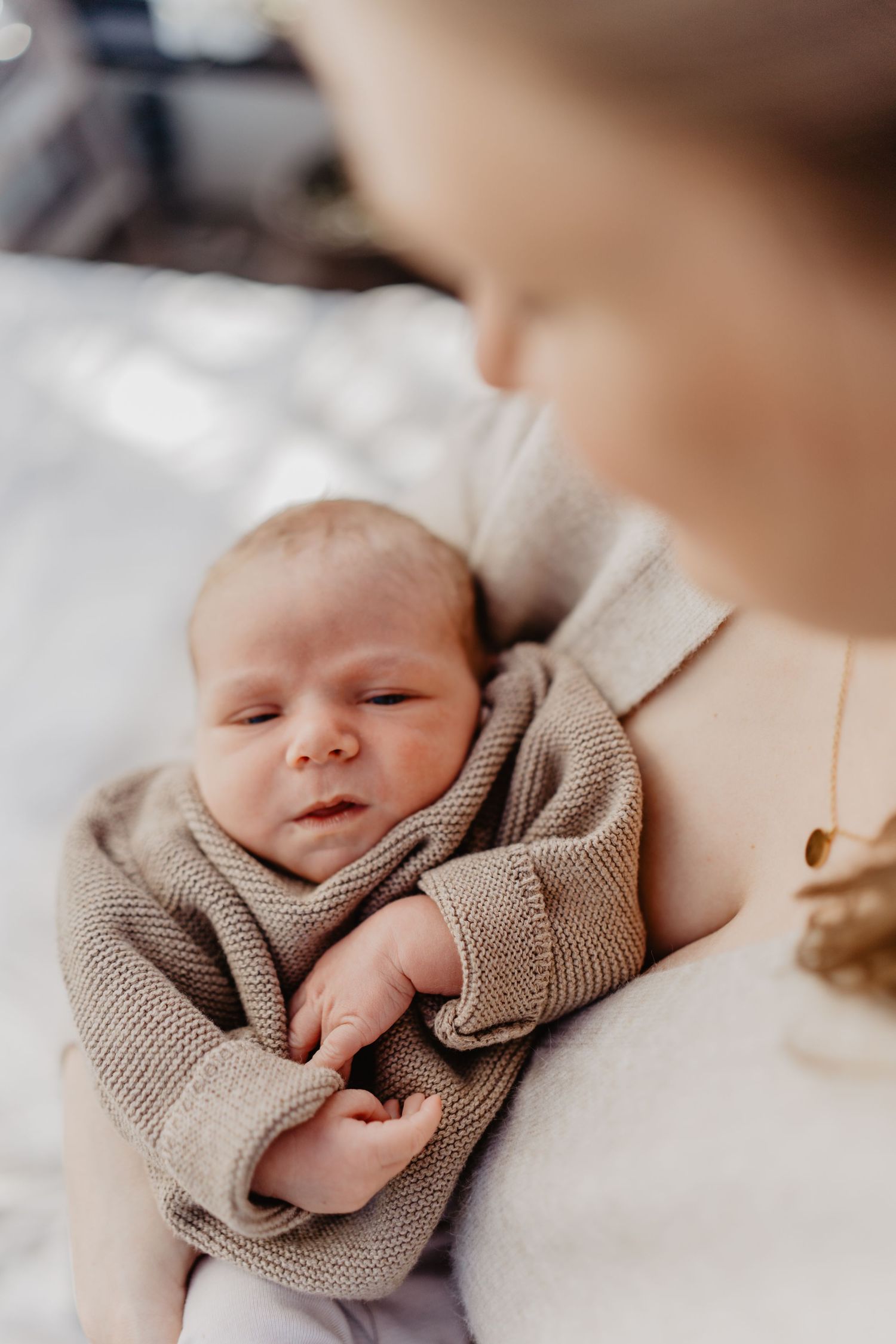 Peaceful newborn wearing cozy beige knit sweater rests on soft white bedding.